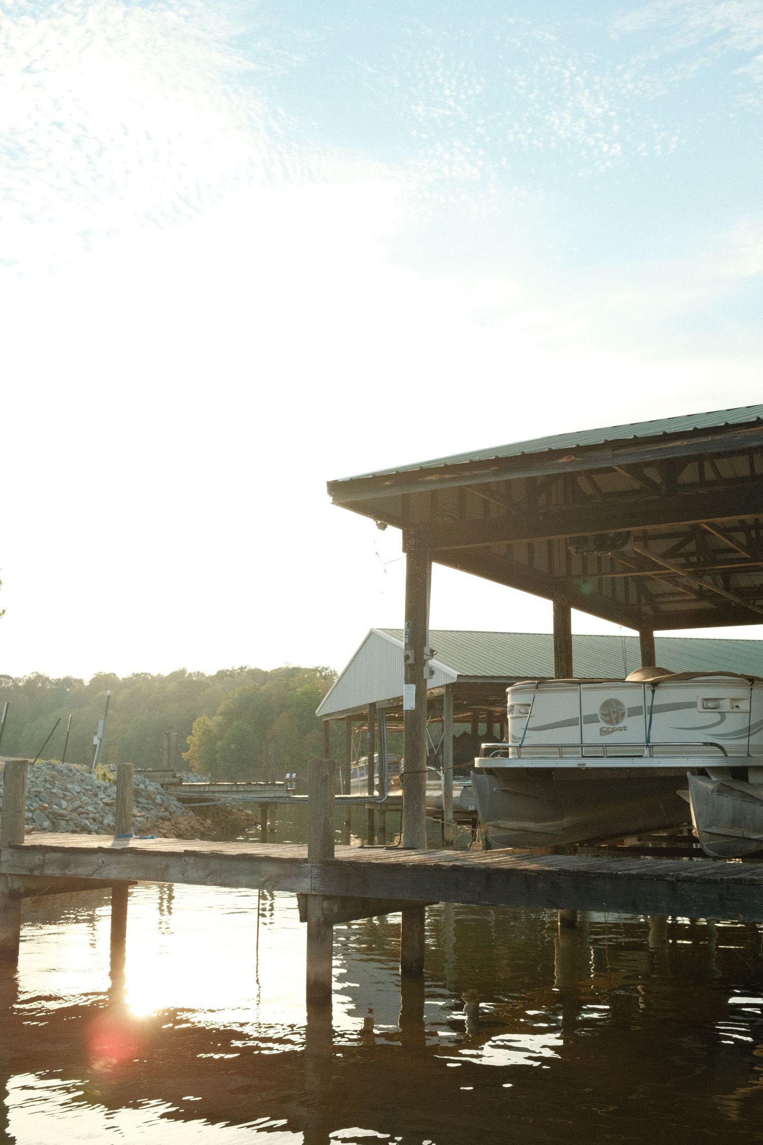 A wooden dock with several boats moored on a calm lake surrounded by trees on a sunny day.