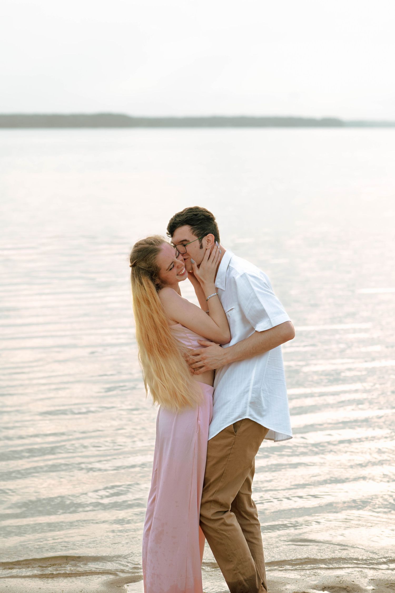 A couple in light summer clothing embraces on a beach at sunset.