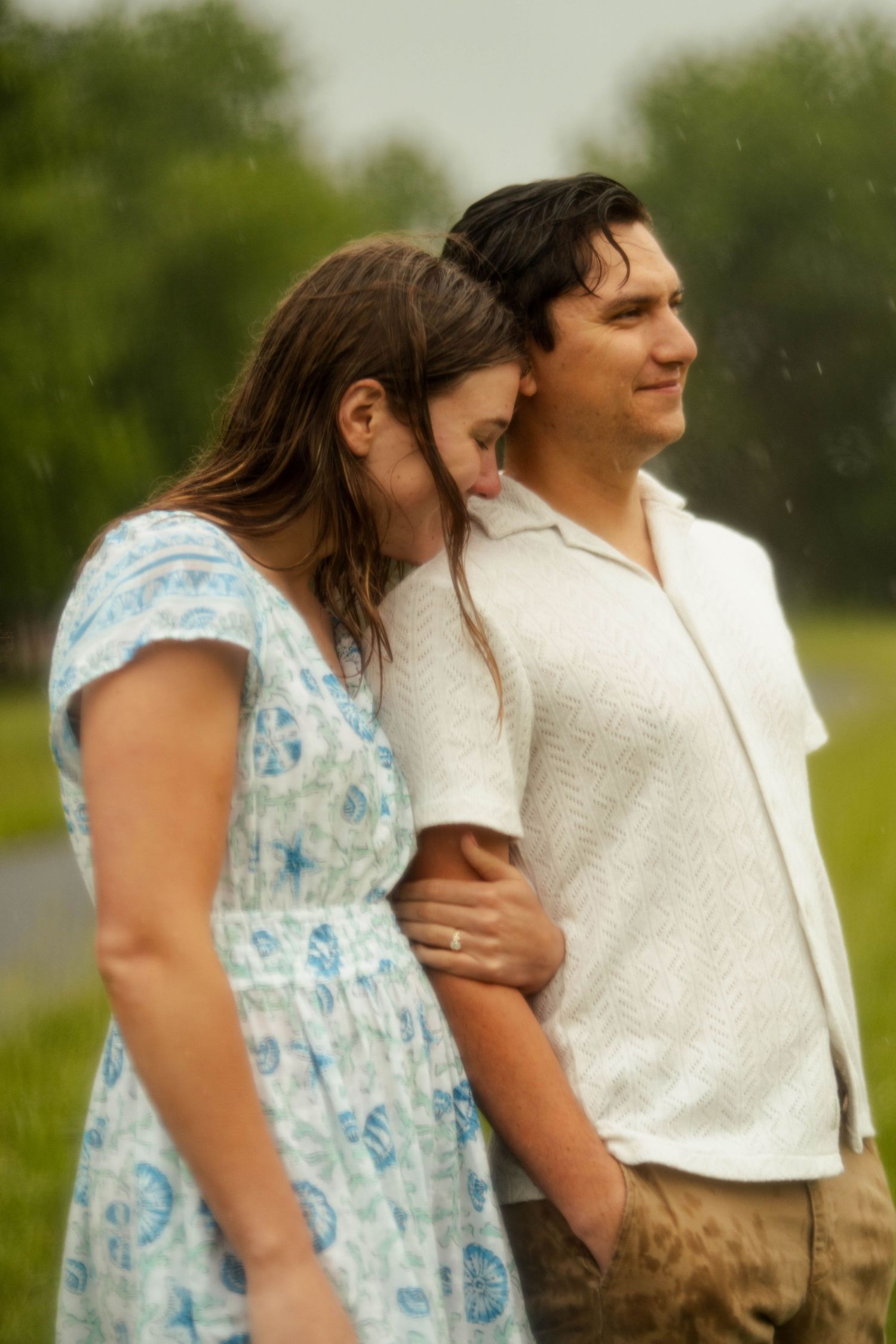 A couple stands together in the rain, woman in floral dress leaning on man in white shirt, outdoors in green field.