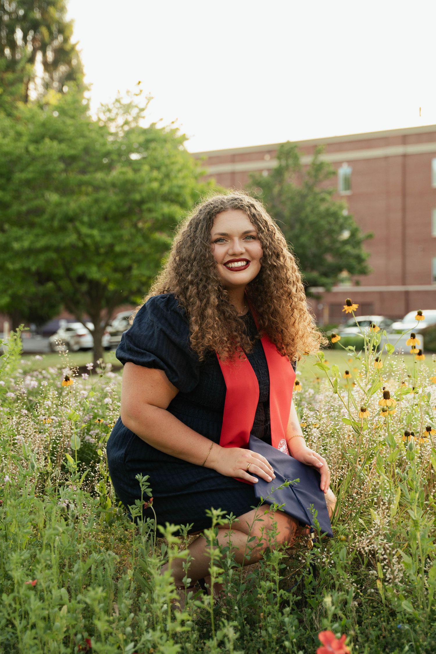 Young woman with curly hair in graduation attire smiling among wildflowers outside a brick building.