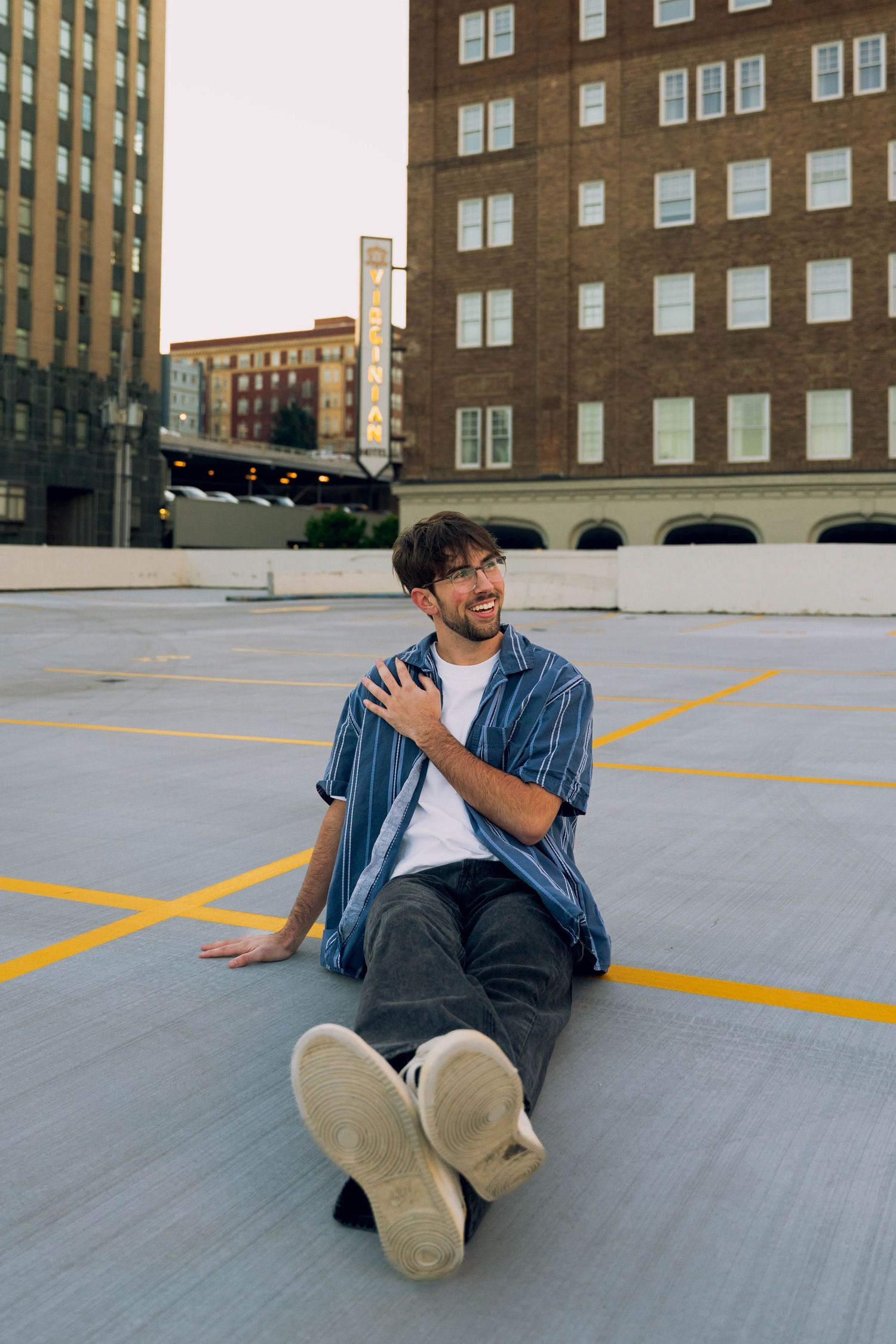 A man wearing a denim shirt and jeans sits on the ground in an urban setting with buildings in the background.