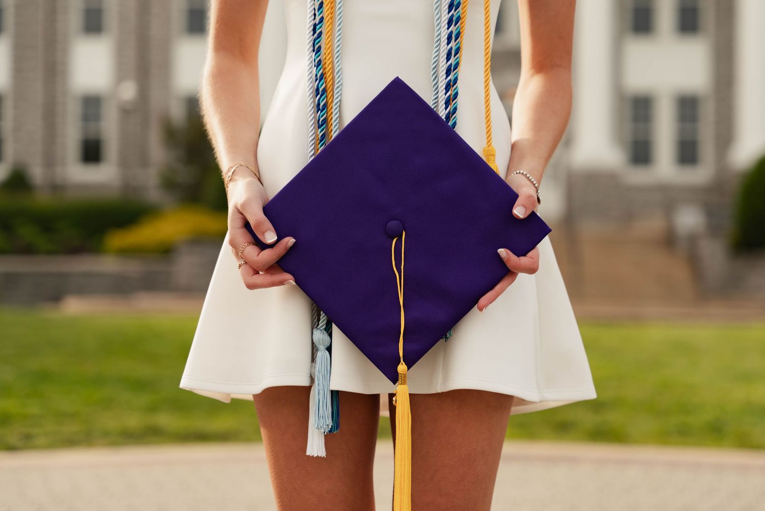 Graduate in white dress holding purple mortarboard cap with gold tassel at college campus ceremony.