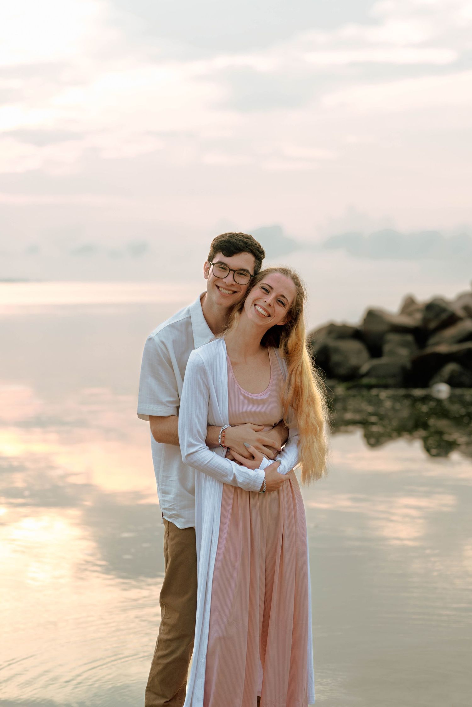 A couple embraces on a beach at sunset with calm waters and rocks in the background.