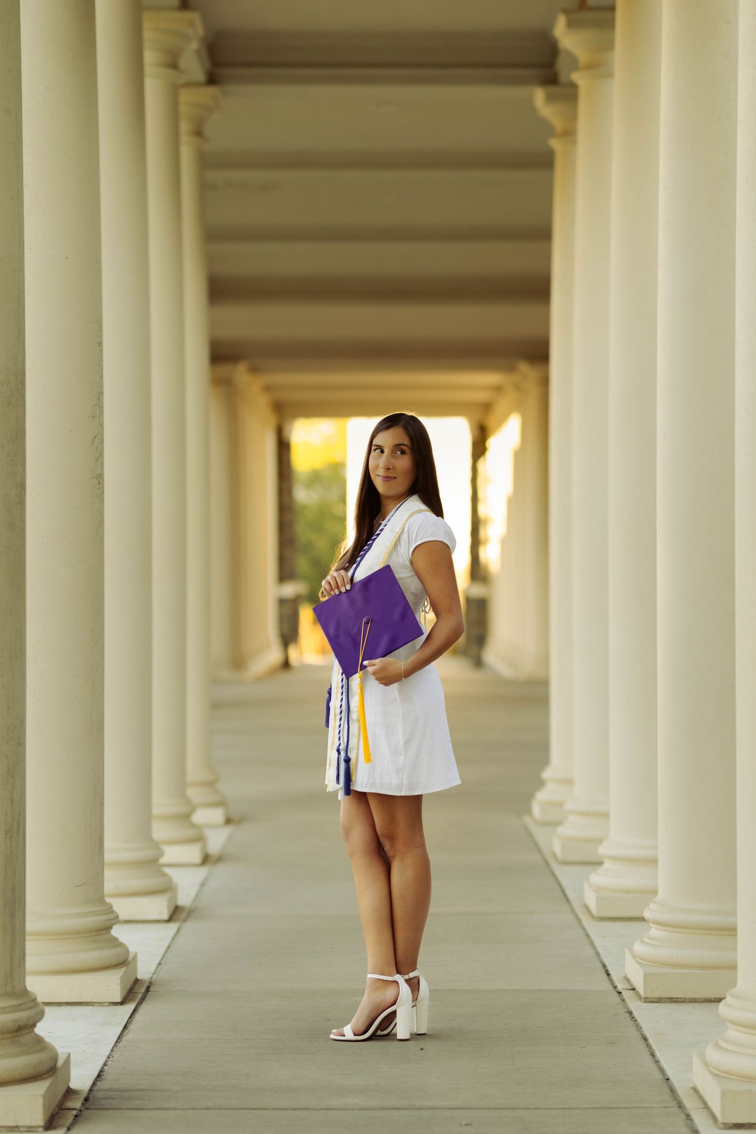 Graduate in white dress poses between classical white columns at university.