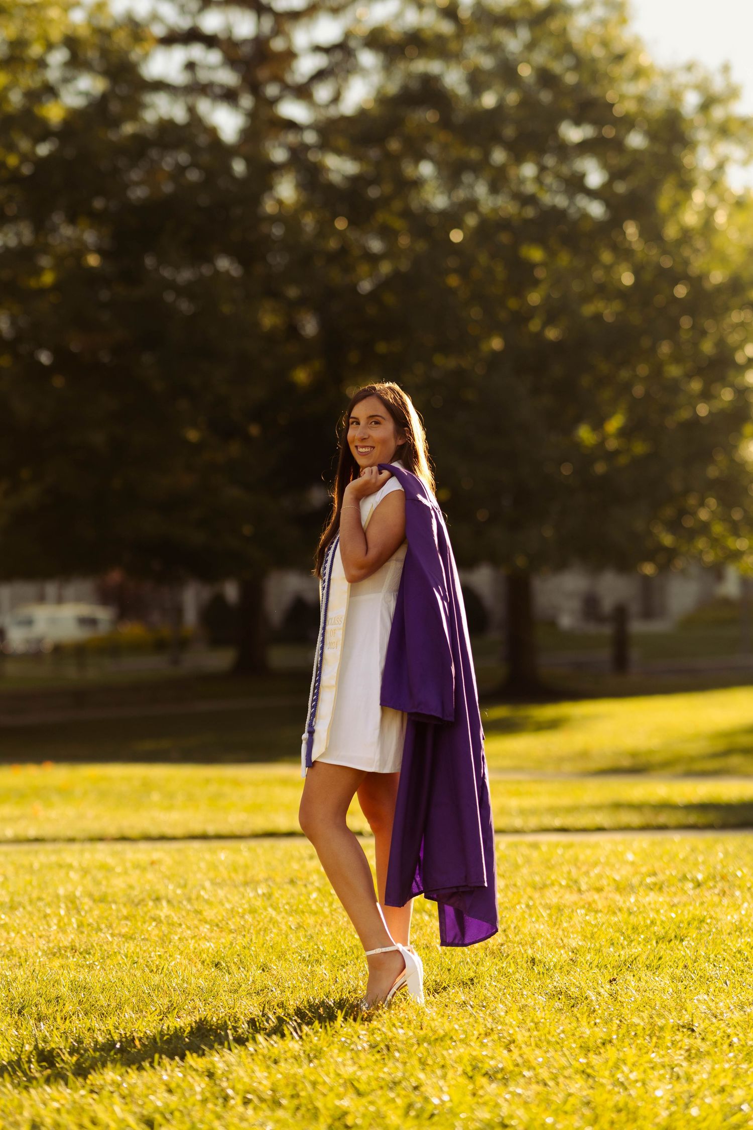 Graduate in white dress and purple stole poses on sunny grass field.