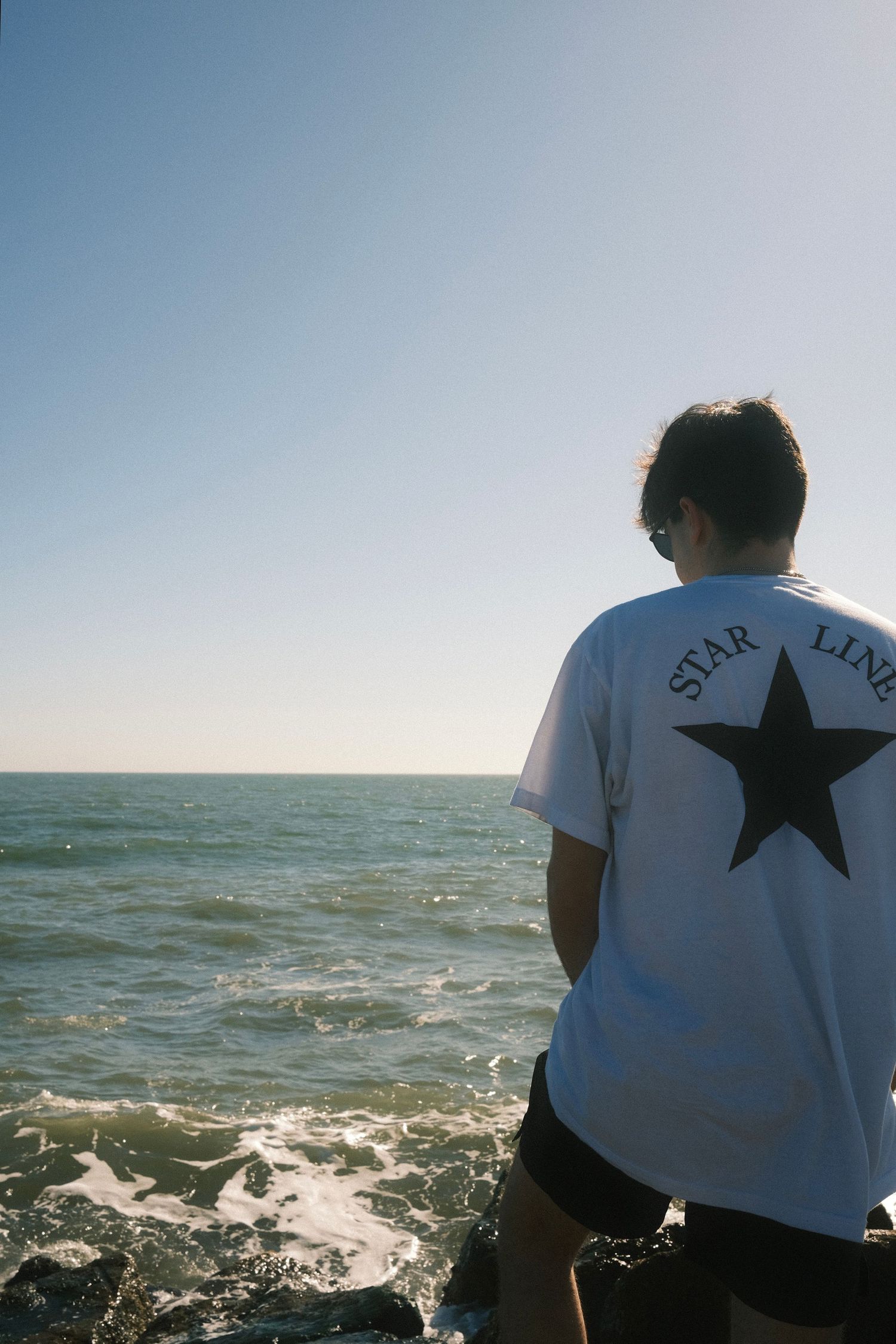 A person wearing a white t-shirt with a star logo stands on a rocky shore overlooking the ocean.