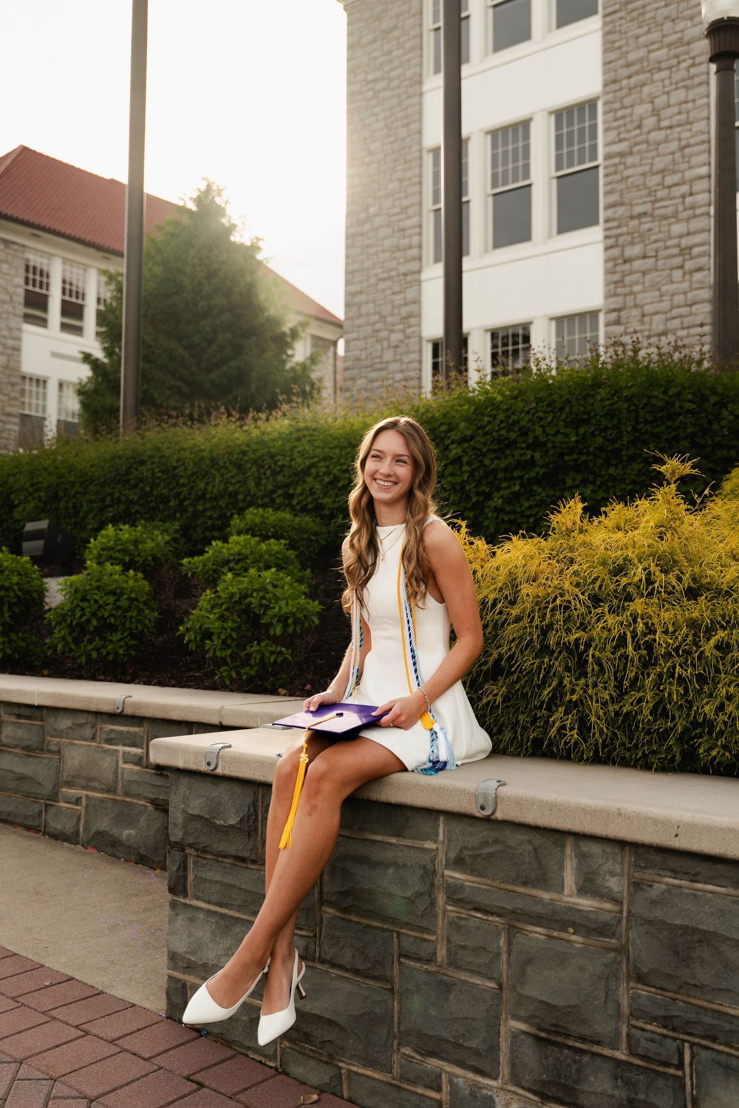Smiling graduate in white dress holding cap and tassel, sitting on stone wall outside college building.