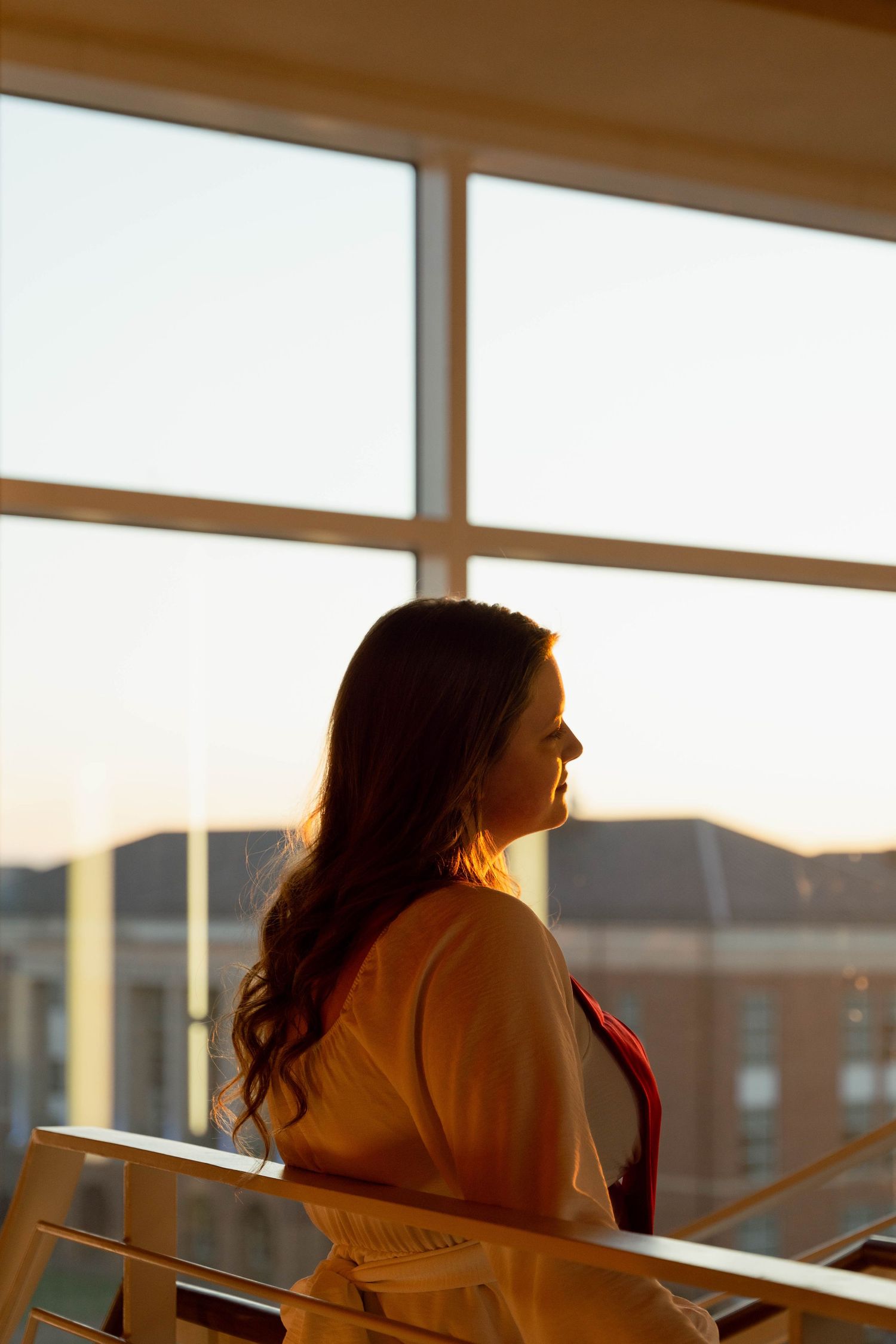 Graduate sits by window during golden hour sunset at university building.