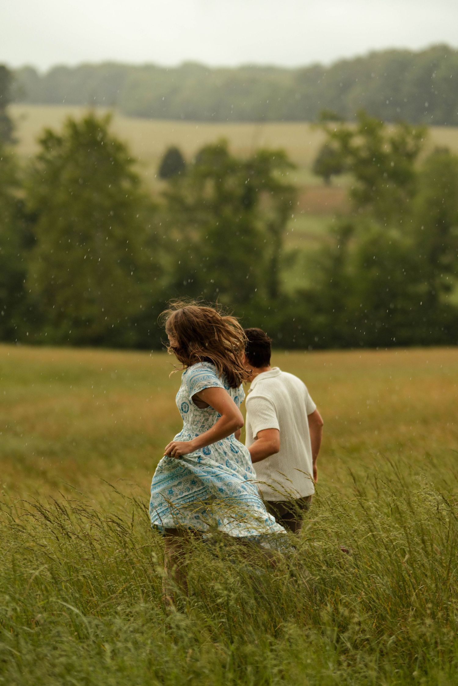 A couple runs through a lush green field in the rain, woman in blue floral dress with hair flowing.