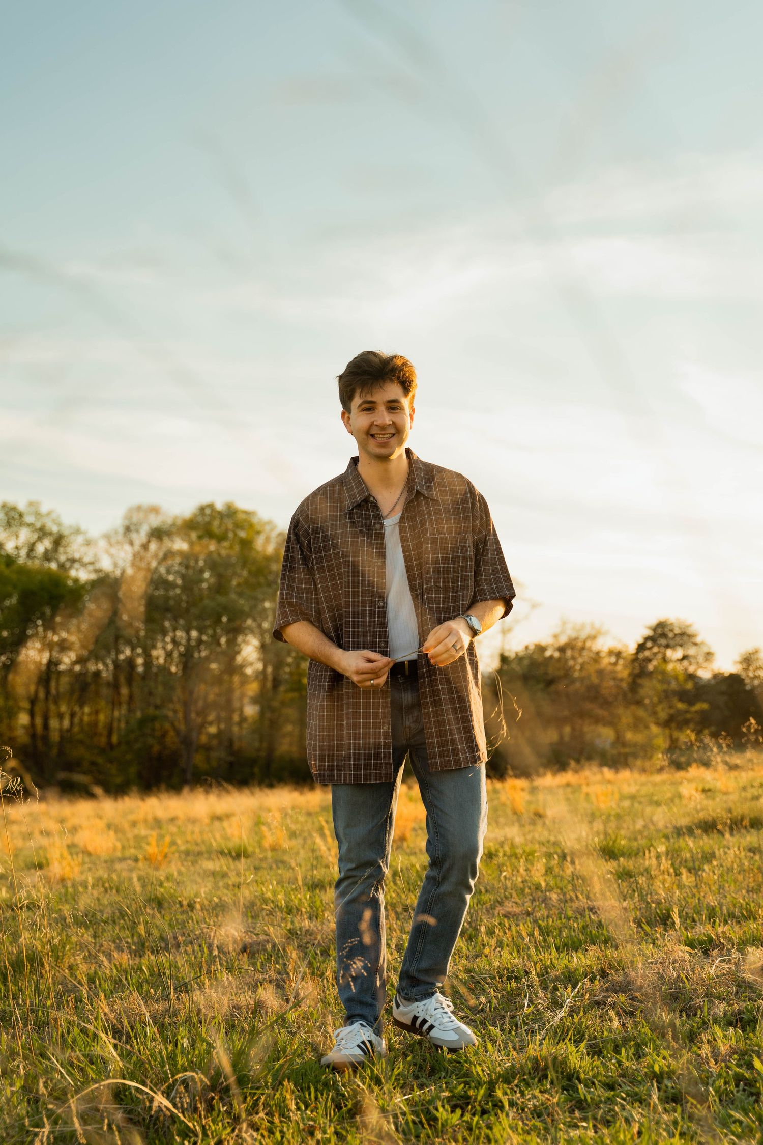 Young man in plaid shirt and jeans smiling in a sunlit field during golden hour.
