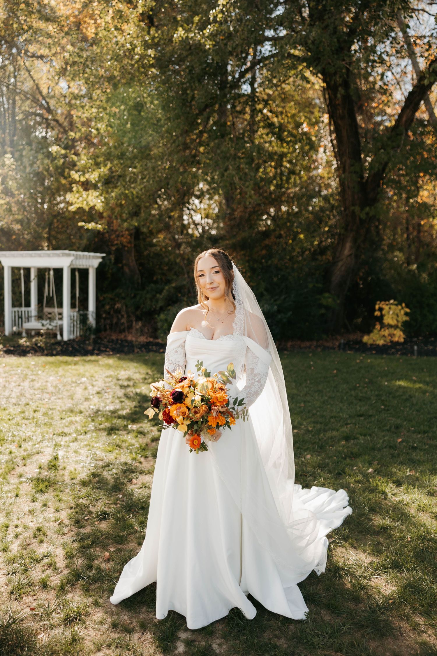 Bride in white off-shoulder gown holding orange fall bouquet, standing on green lawn with gazebo and autumn trees.