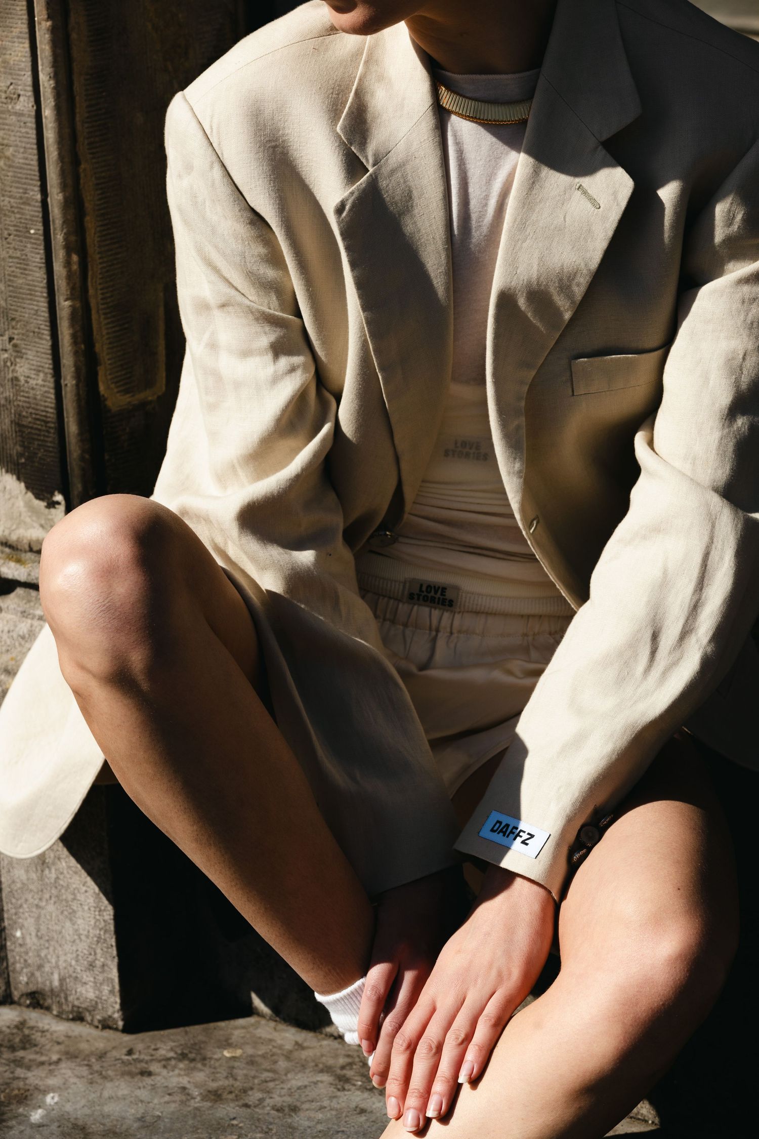 Model in beige linen blazer and shorts seated outdoors in sunlight with wristband detail.