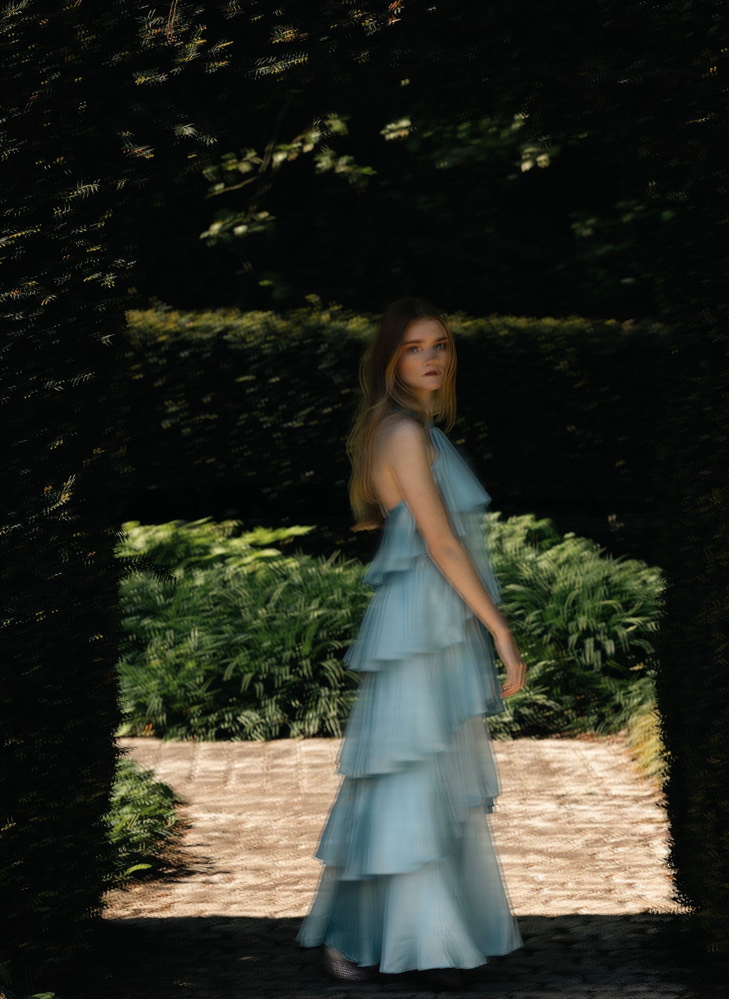 Woman in tiered light blue ruffle gown stands in elegant garden archway surrounded by lush hedges.