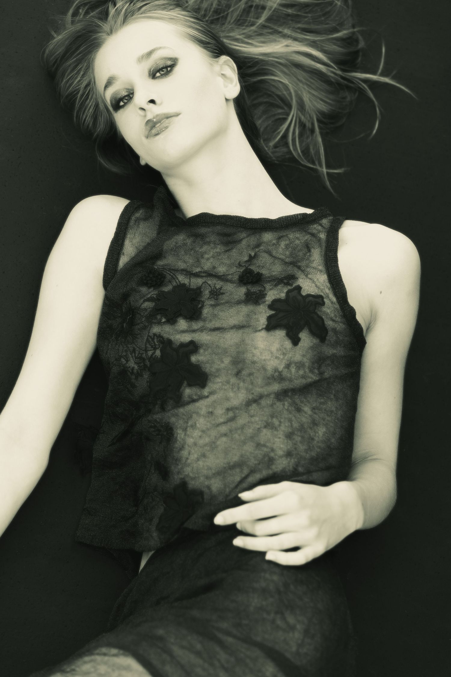 Black and white photo of a woman lying down wearing a sheer floral dress with windswept hair.