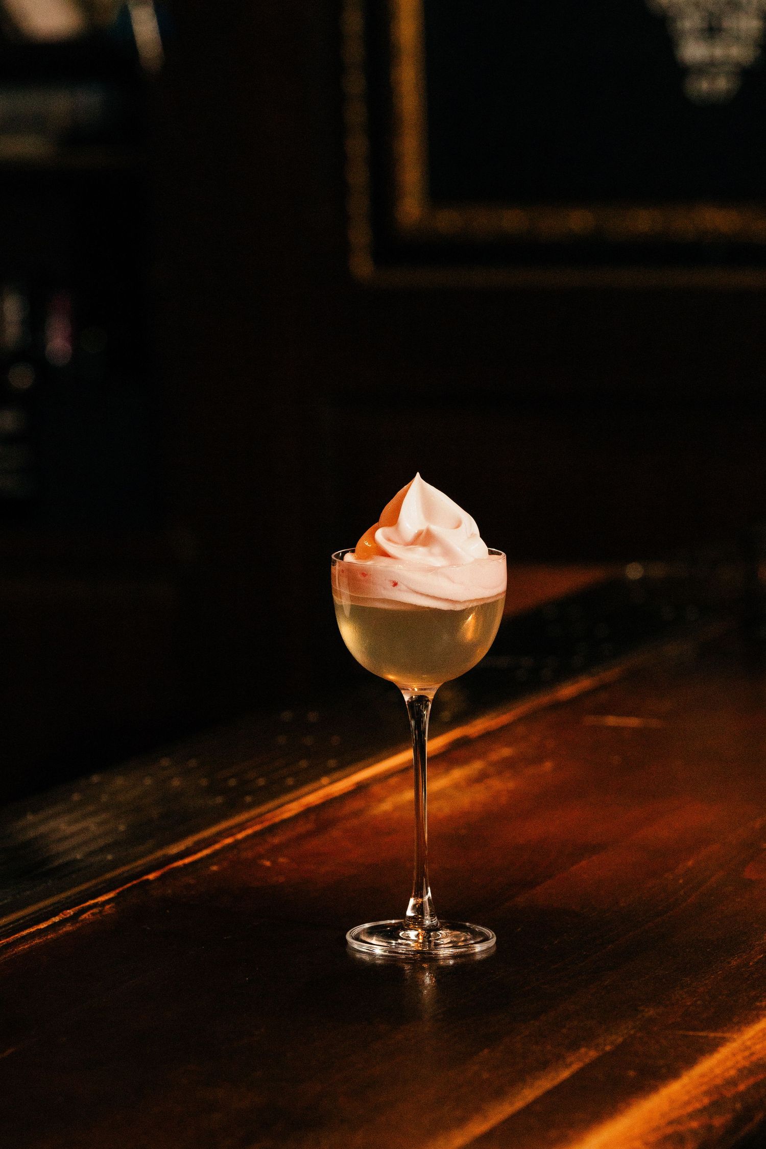 Sophisticated cocktail topped with swirled pink whipped cream in stemmed glass on dark bar.