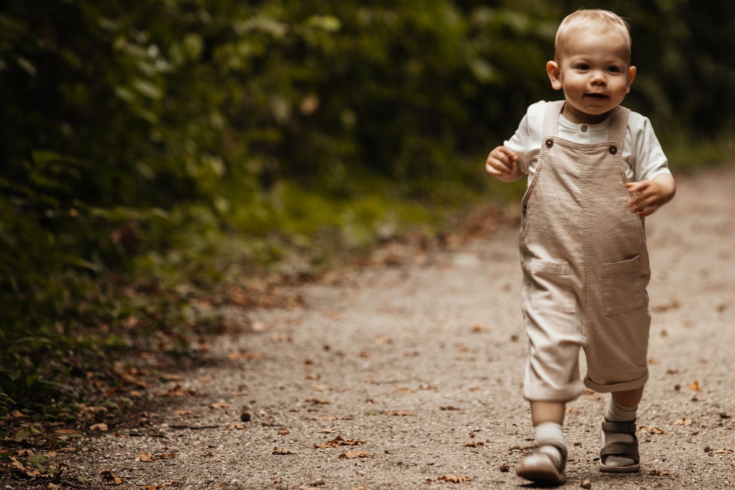 A cute toddler in beige overalls walking confidently along a garden path surrounded by lush greenery.