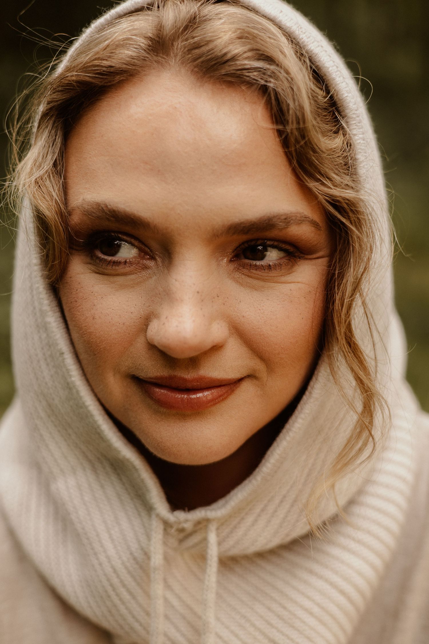 Close-up portrait of a young woman with blonde hair wearing a cream hoodie, smiling softly outdoors.