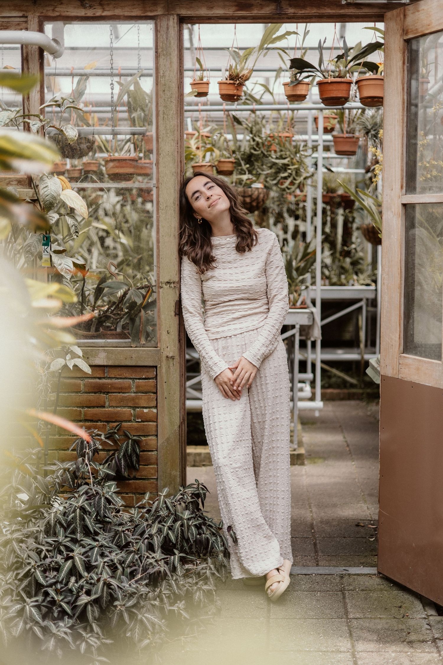 Woman in cream knit outfit leaning against greenhouse door surrounded by lush green plants.