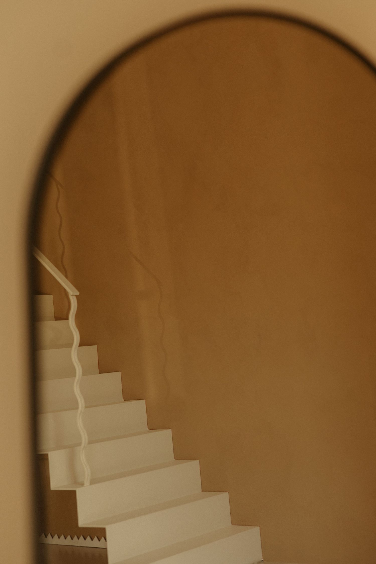 Arched mirror reflects a white staircase with railing against a warm golden-beige wall.