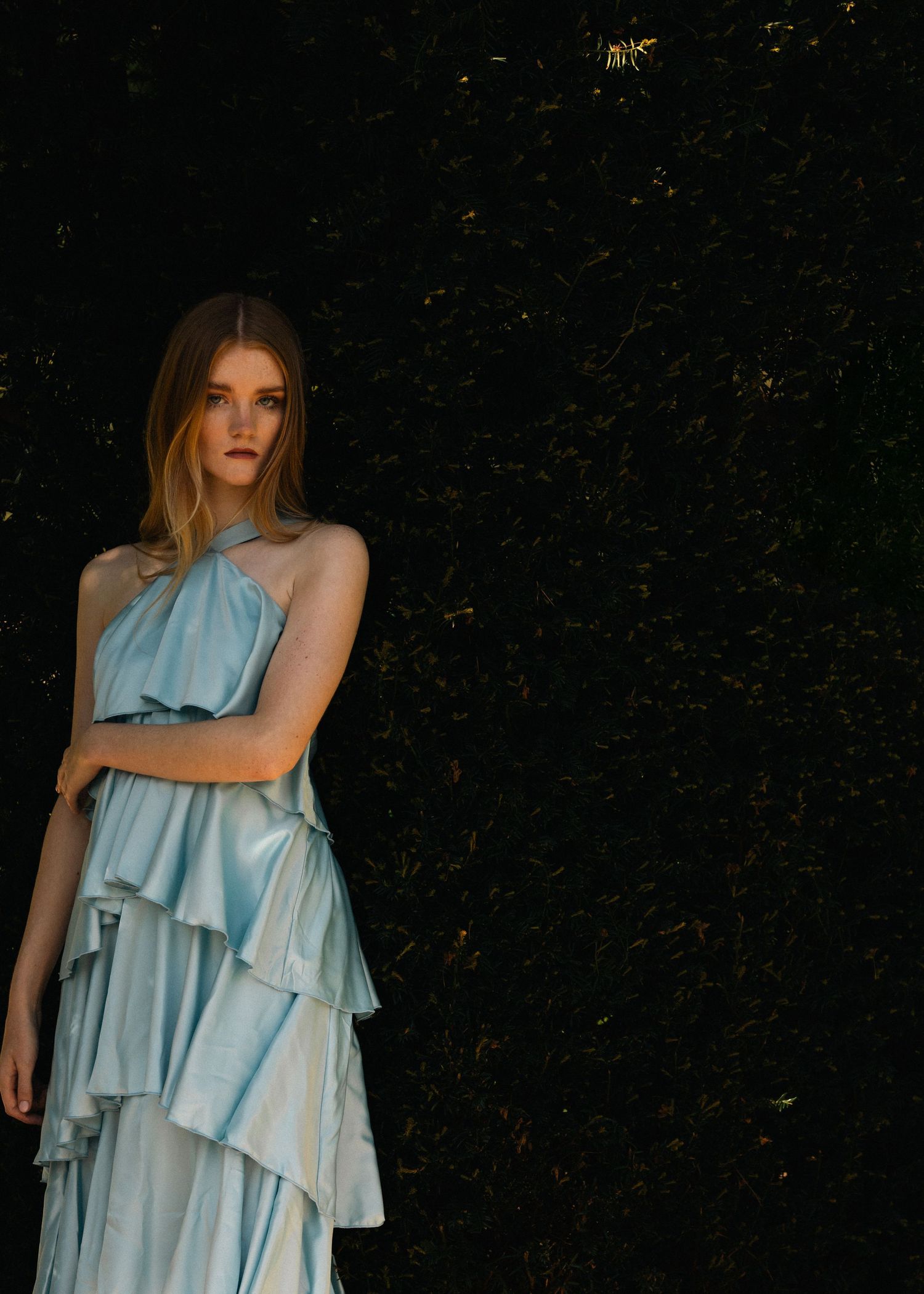 Woman in light blue ruffled halter dress standing against dark green hedge backdrop.