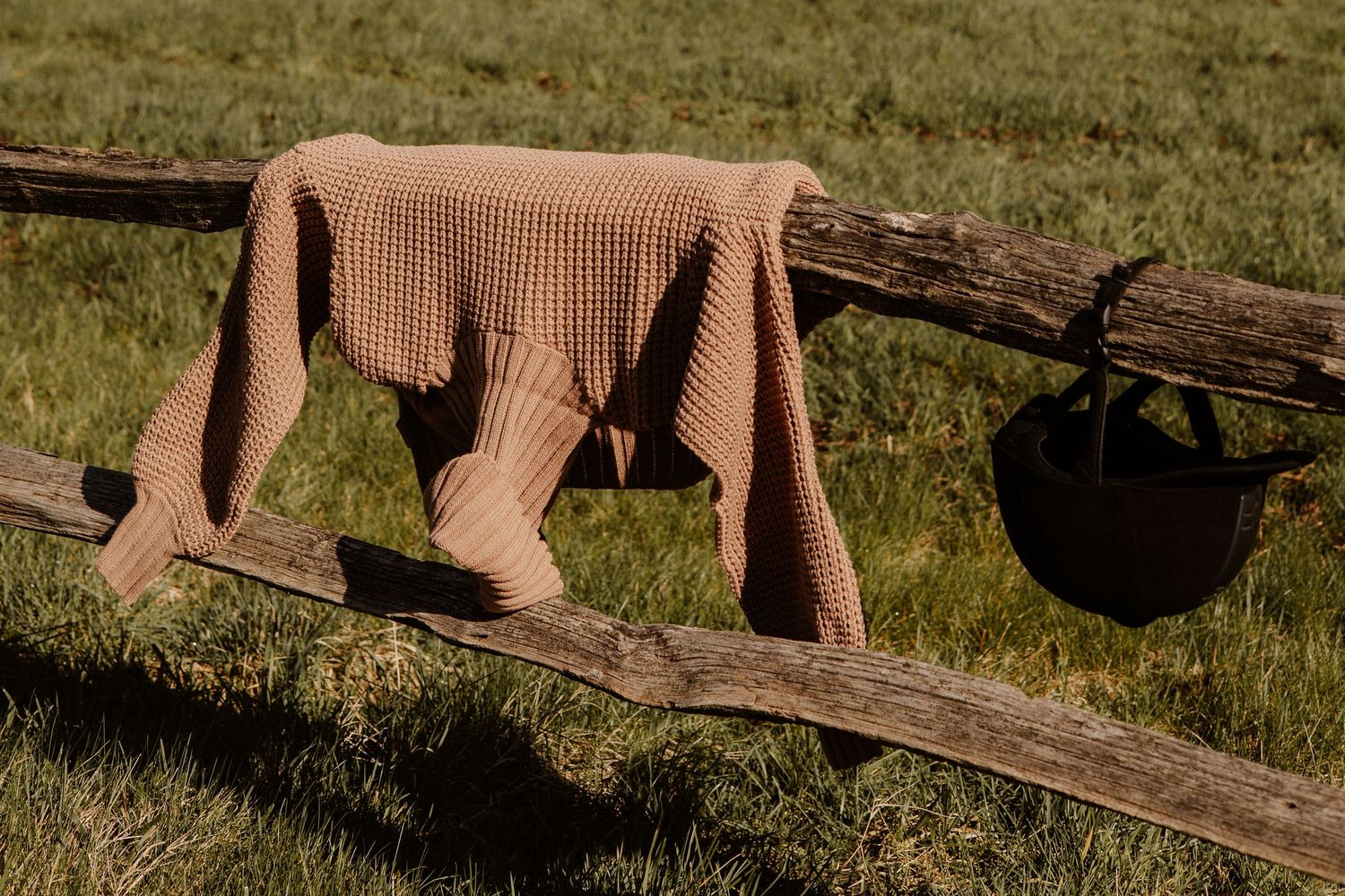 Brown waffle-knit sweater and black hat draped over a rustic wooden fence in a sunny field.