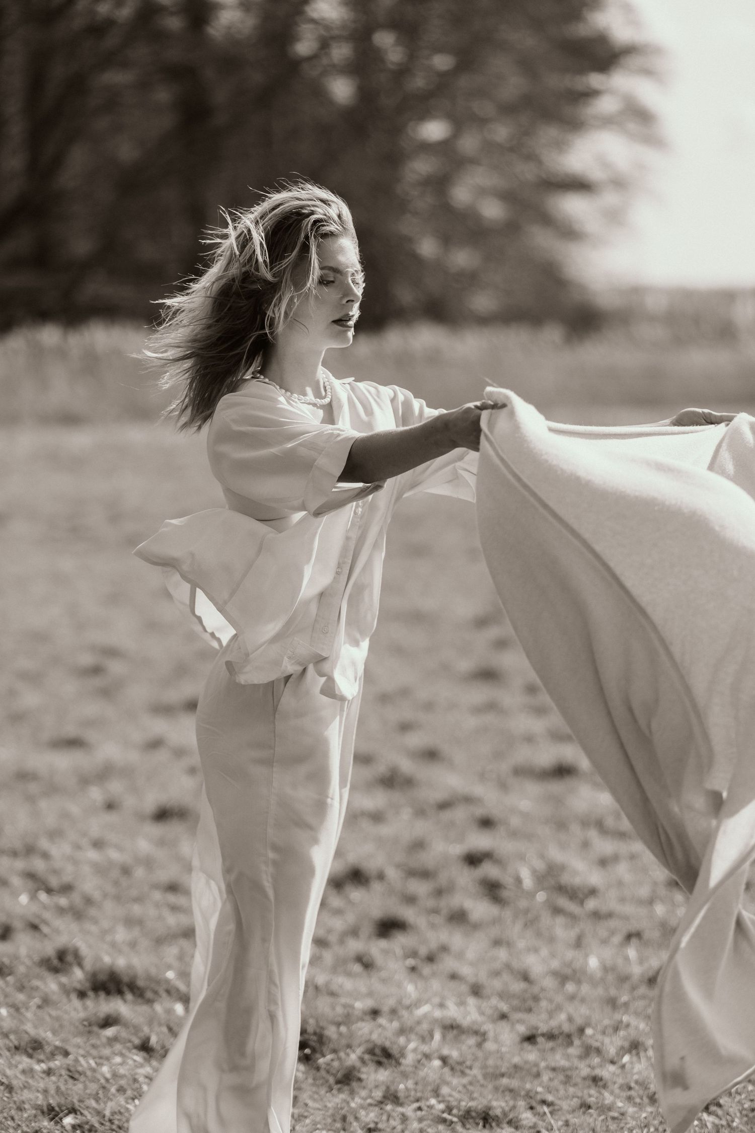 Black and white photo of a woman in white linen outfit holding flowing fabric in an open field.