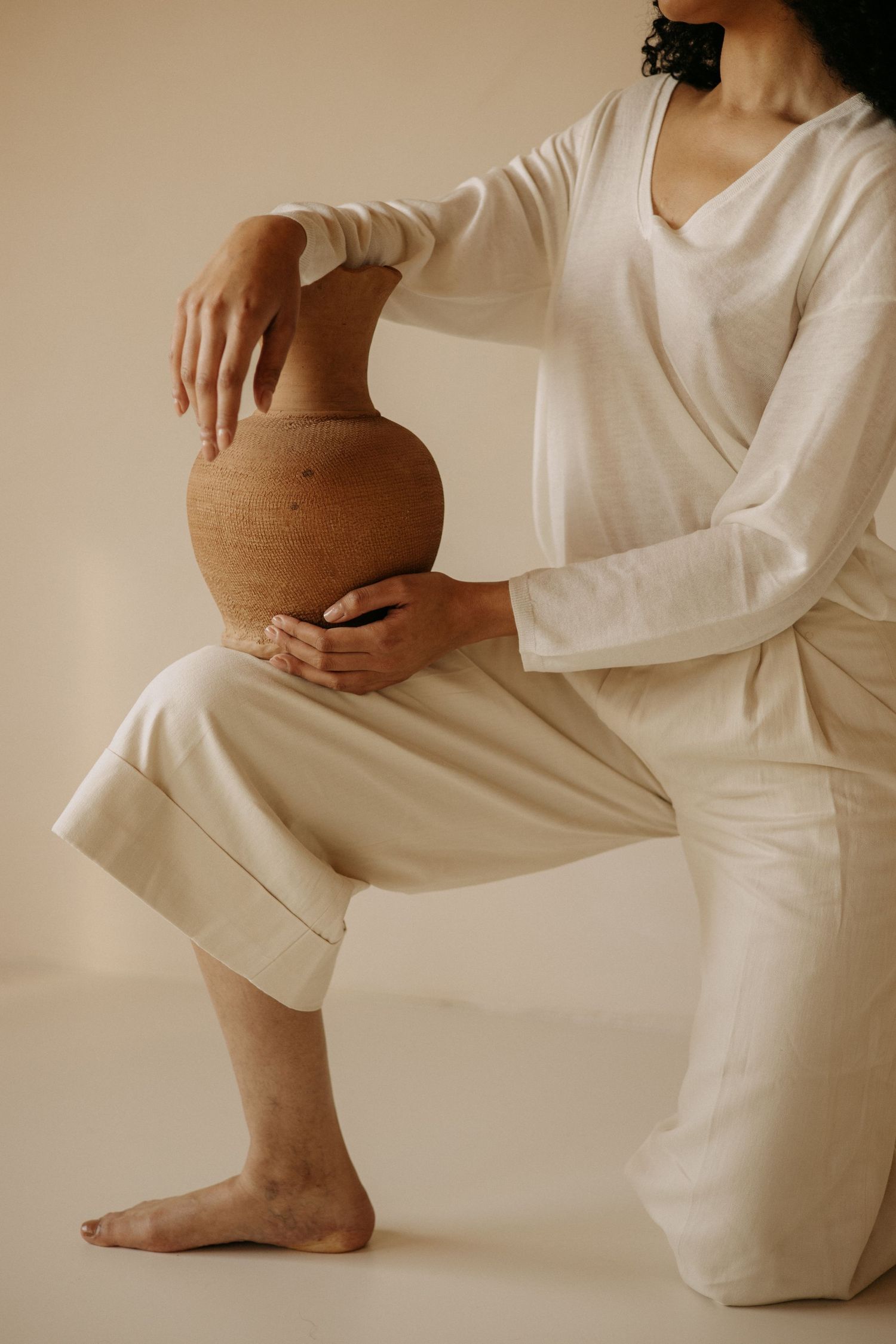 Person in white linen outfit kneels while holding a textured terracotta clay vase.