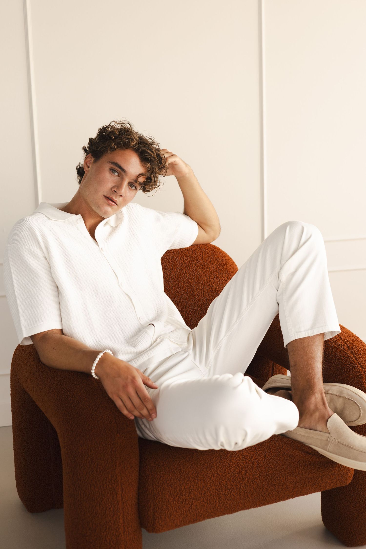 Young man in all-white outfit relaxing on brown textured armchair.