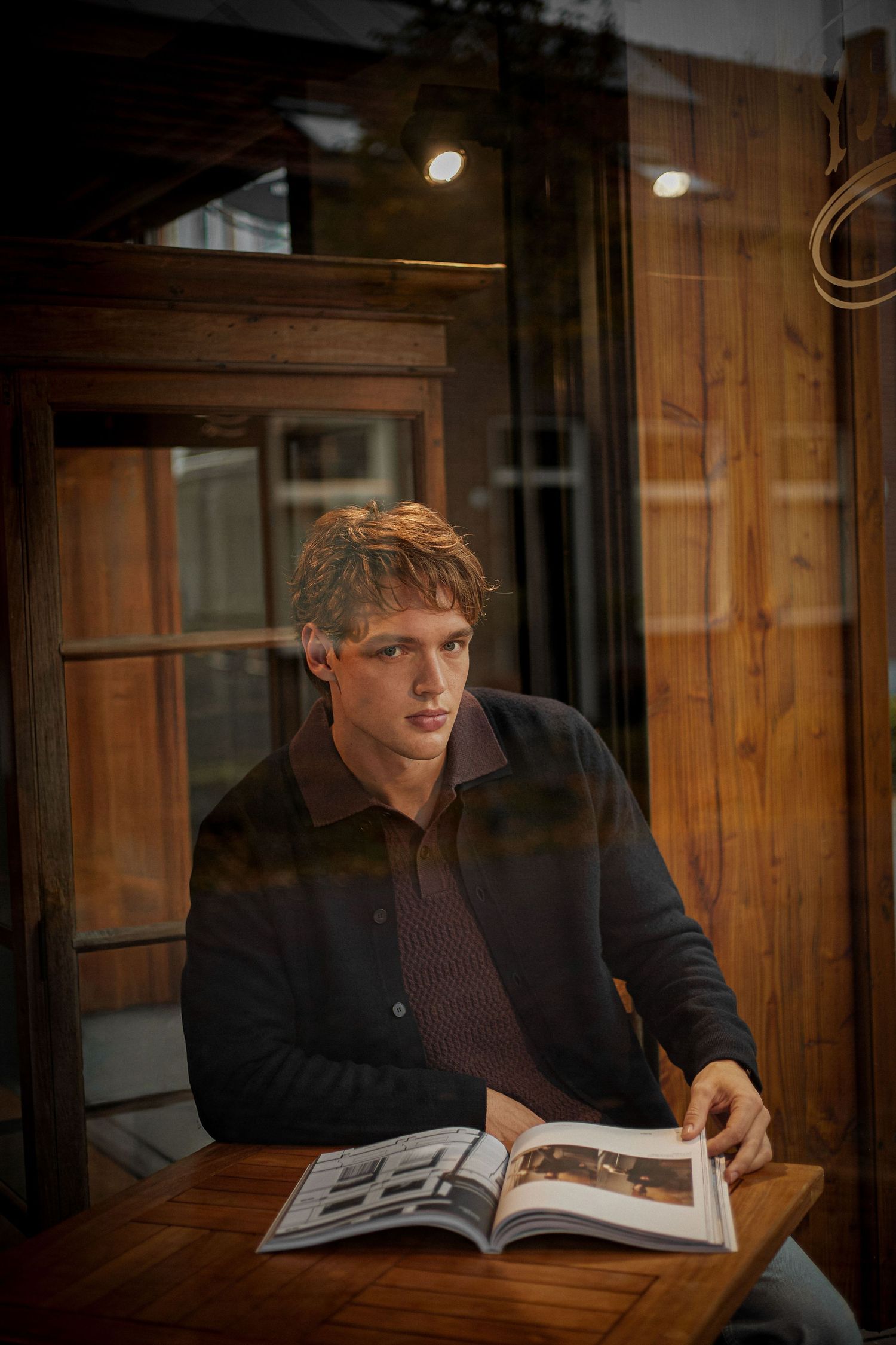 Young blond man reads magazine at wooden table in warmly lit rustic interior setting.