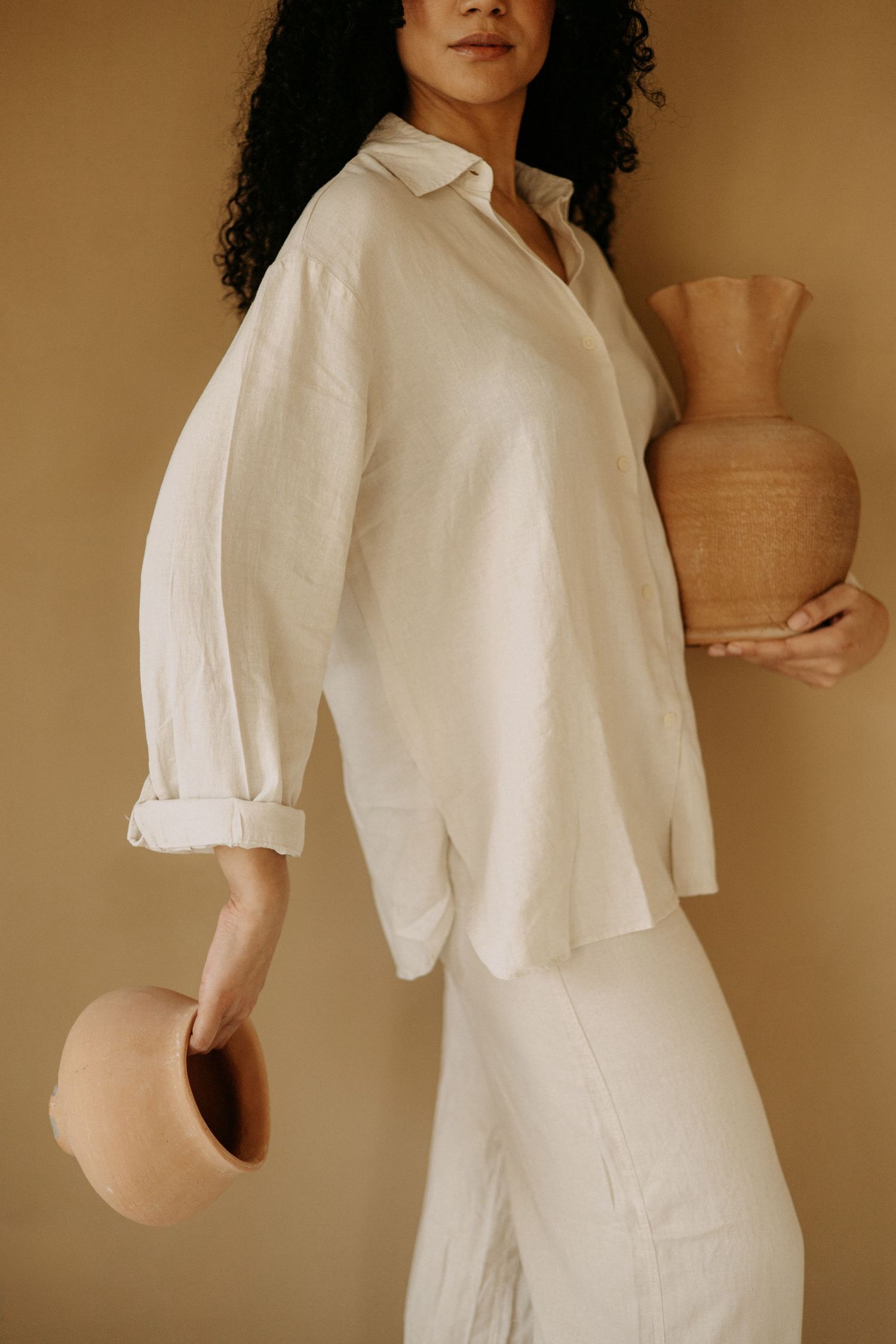Woman with curly hair in white linen shirt holds two terracotta clay vases against a beige backdrop.