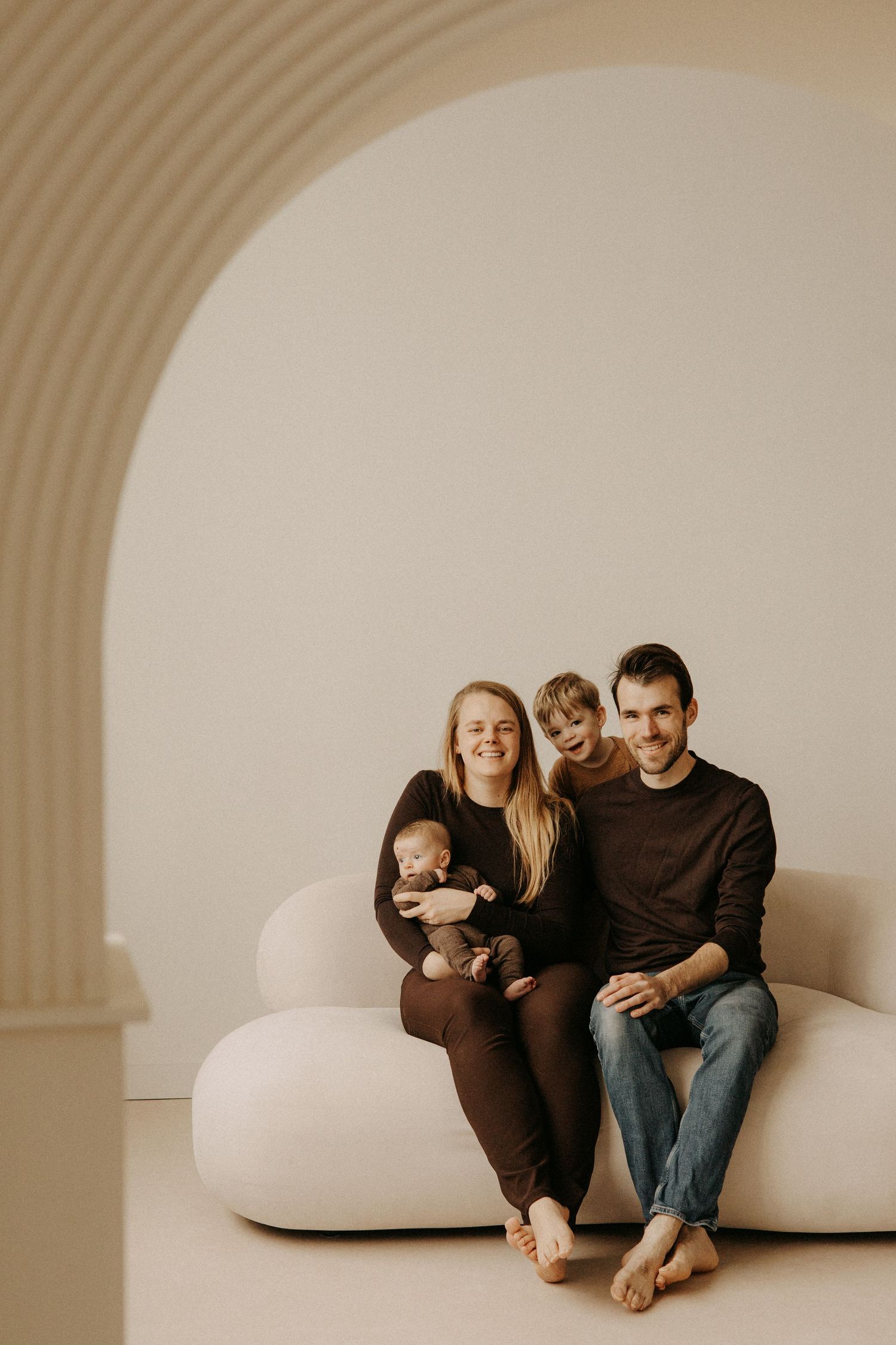 Happy family with newborn baby and toddler posing on white curved sofa in minimalist beige studio.