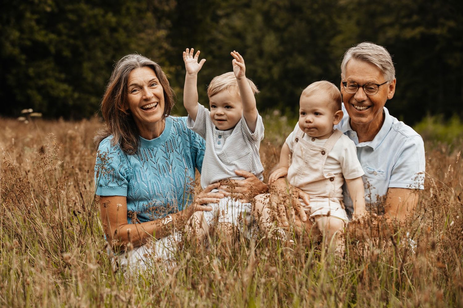 Laughing grandparents sitting in a tall grass field with two young grandchildren waving during a family photo.