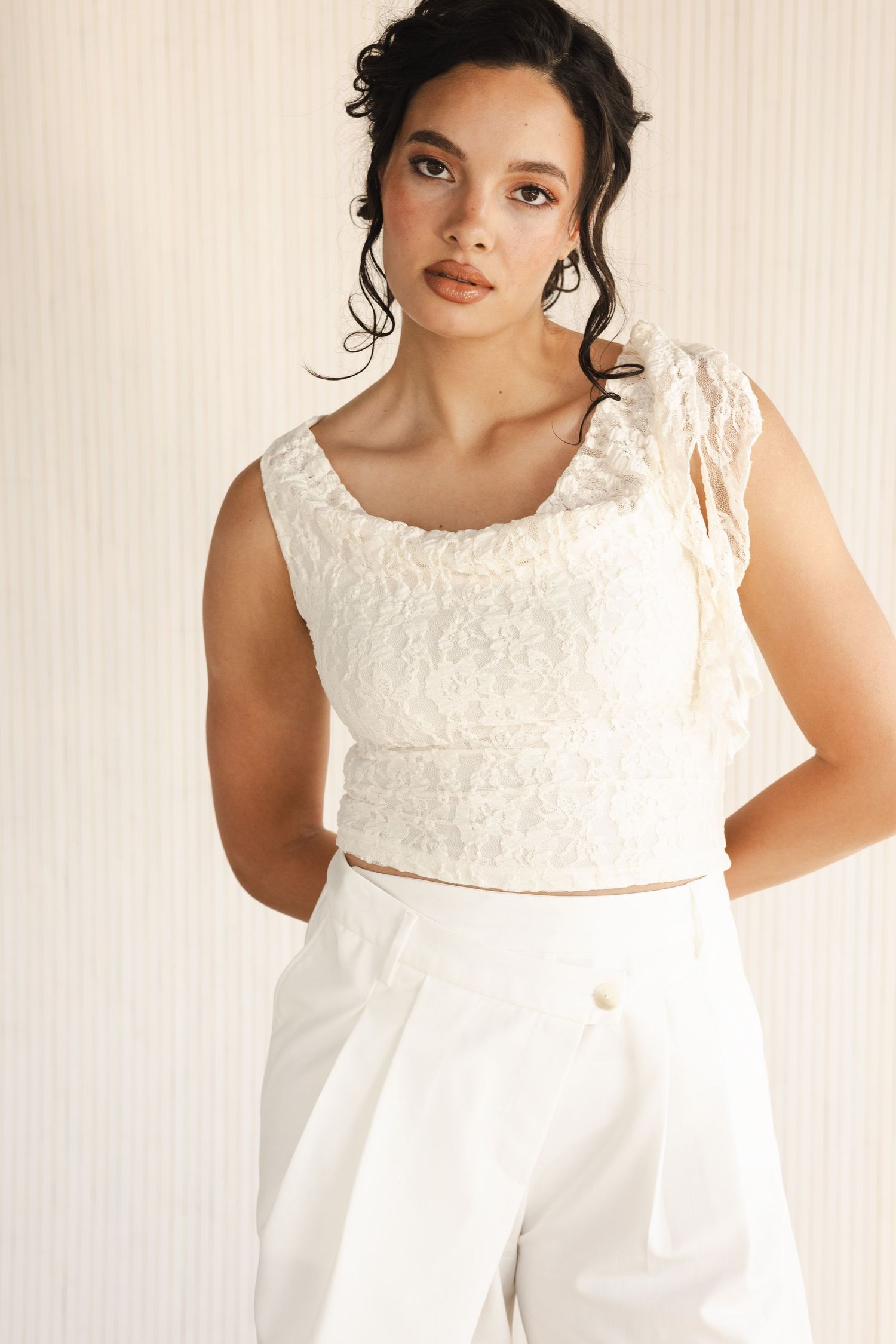 Woman in ivory lace crop top and white wide-leg pants posing against striped background.