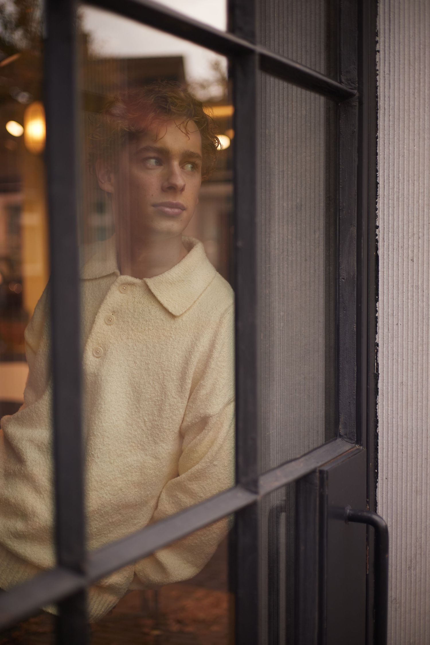 Young man in cream knit polo sweater looking through glass window door.