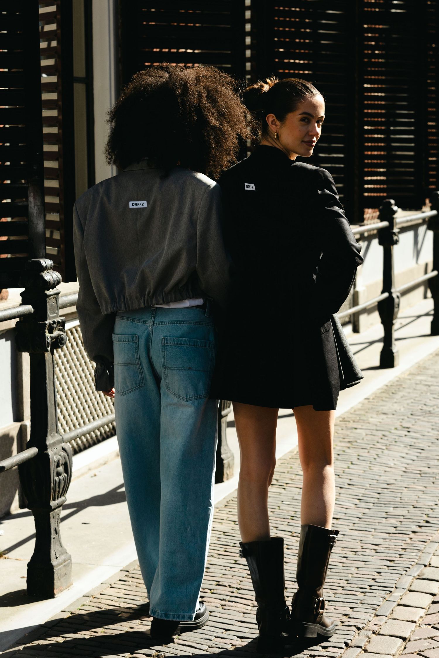 Two women in black jackets stand on a cobblestone street, one in blue jeans, one in a mini skirt with combat boots.
