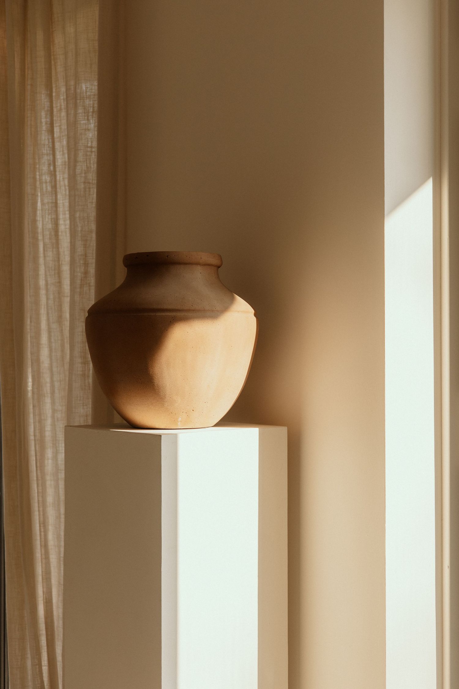 Rustic clay pottery vase displayed on white pedestal in sunlit minimalist interior space.