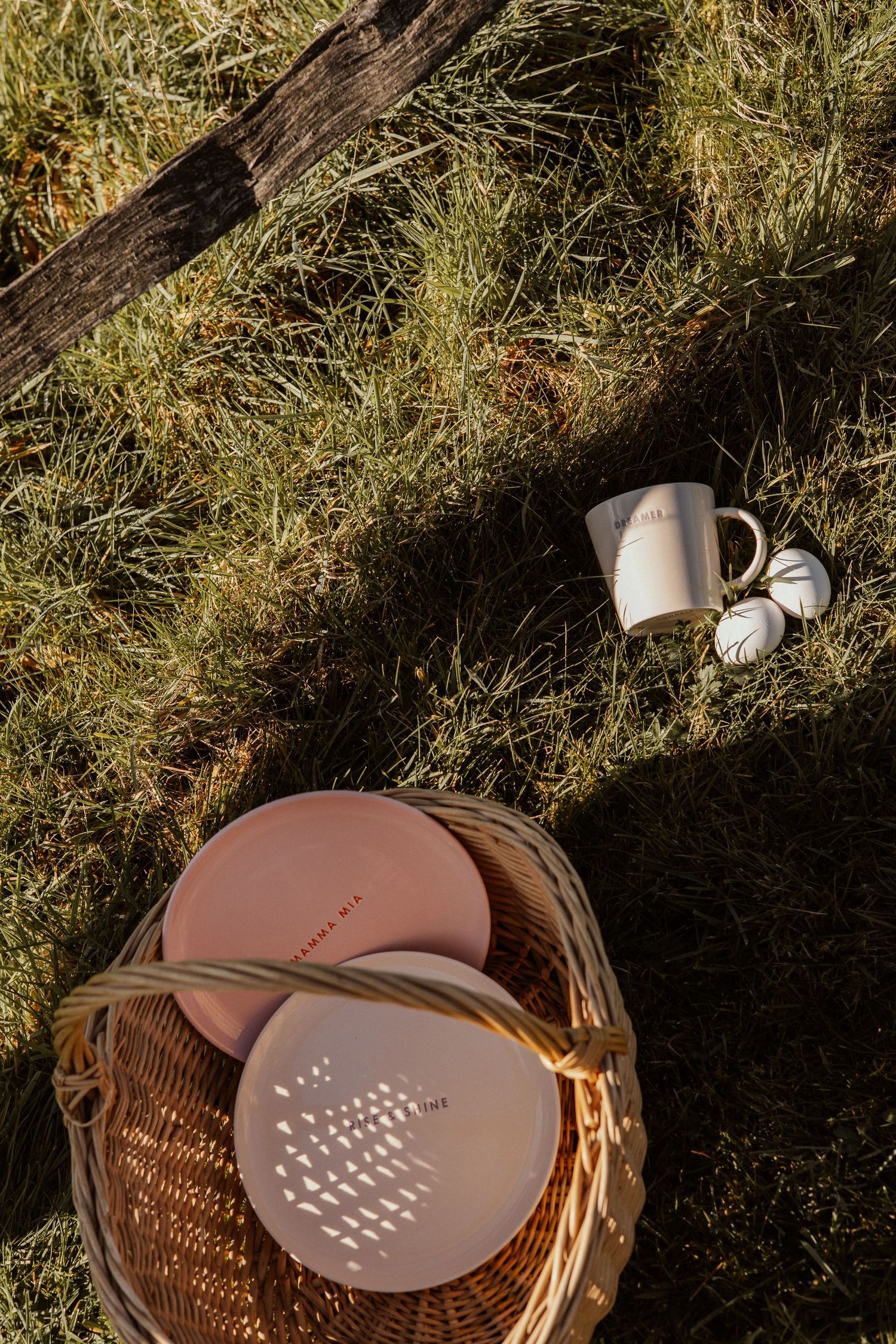 Wicker basket with pink plates and a white mug resting on green grass in warm sunlight.