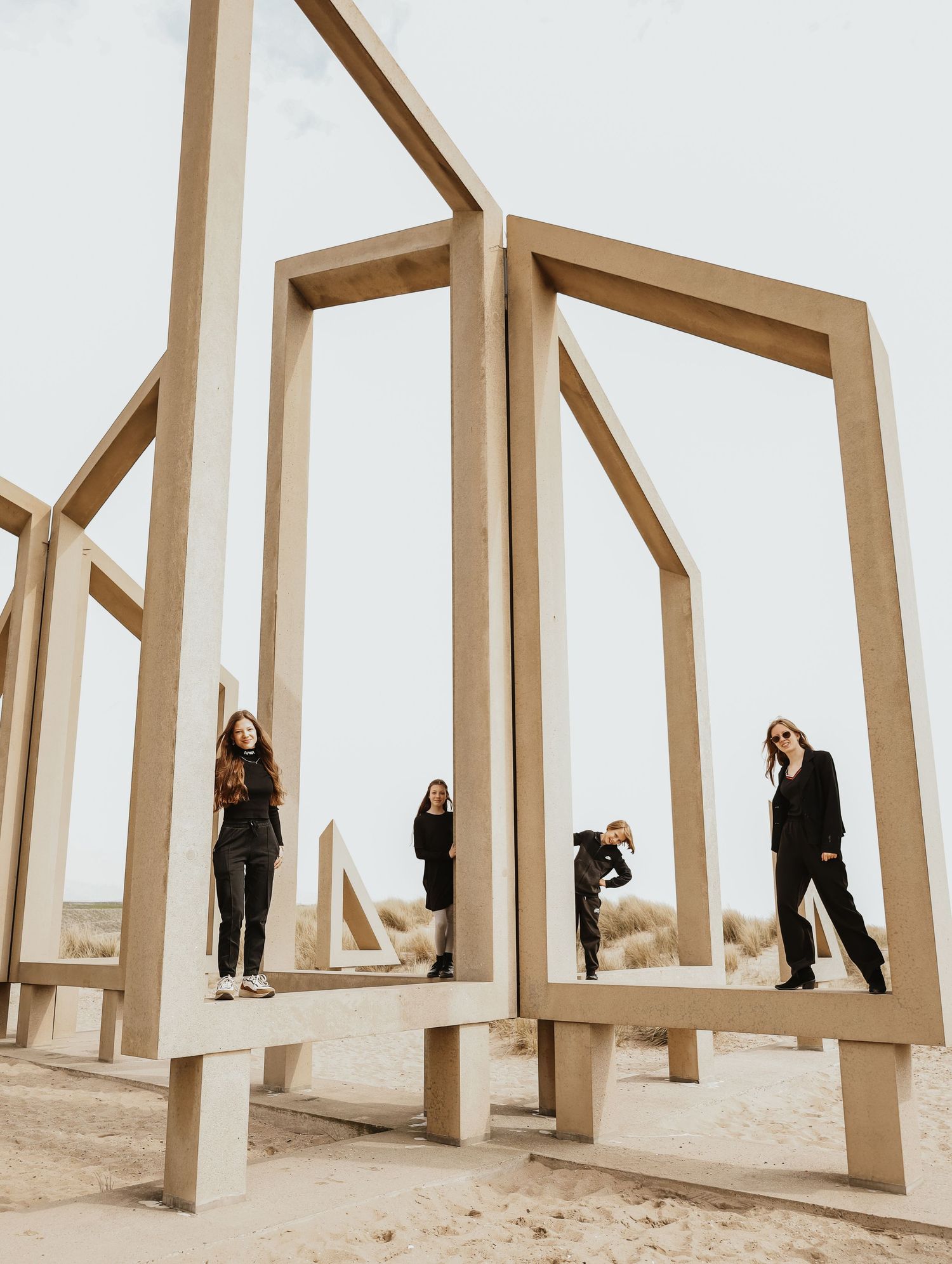 Four people posing among large geometric wooden frame structures on a sandy beach under a cloudy sky.