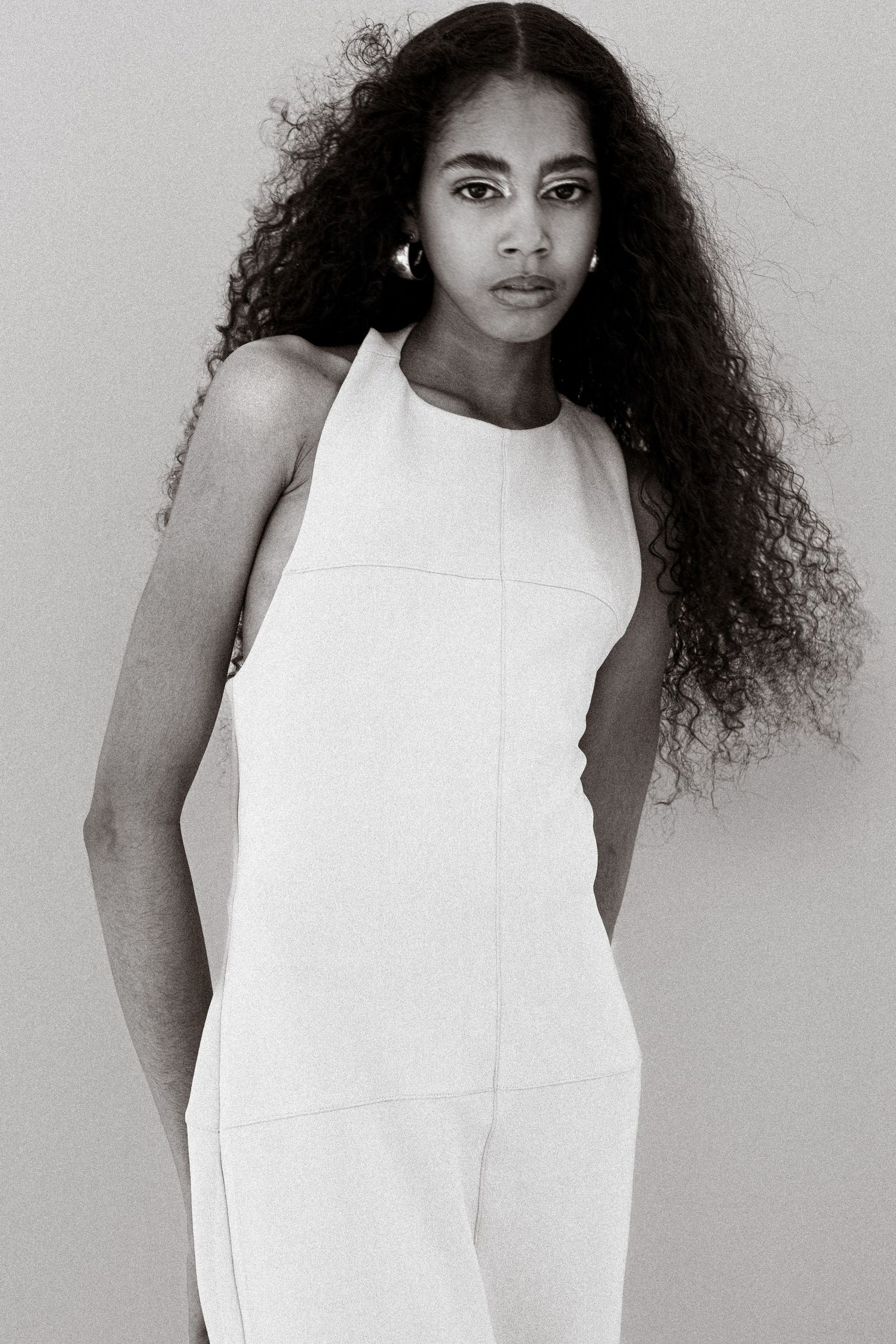 Black and white portrait of young woman with long curly hair in white sleeveless jumpsuit.