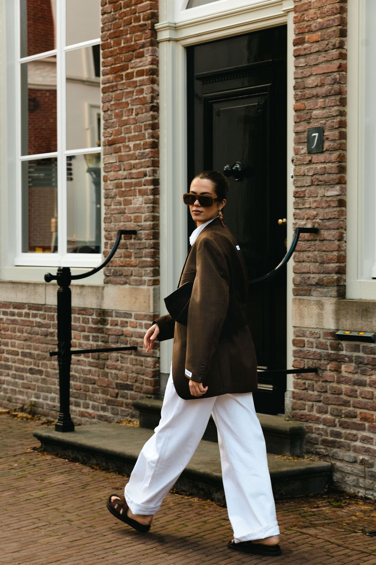 Woman in brown oversized blazer and white wide-leg trousers walks past brick building.