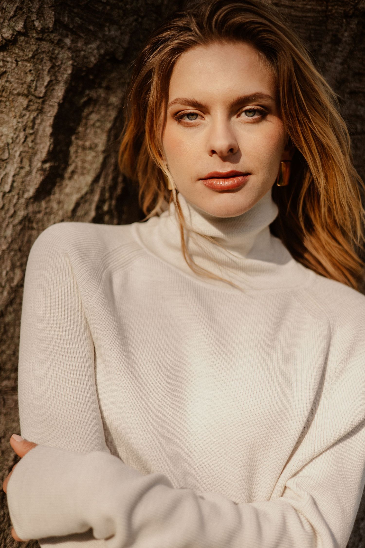 Woman in cream turtleneck sweater leaning against tree trunk in warm golden light.