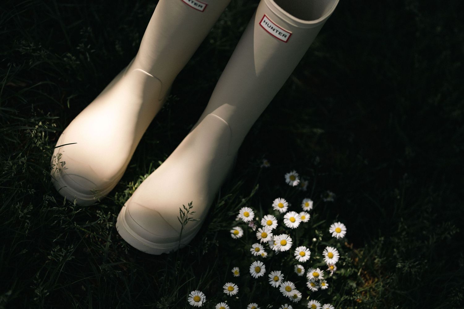White Hunter rain boots beside white daisies on dark grass.