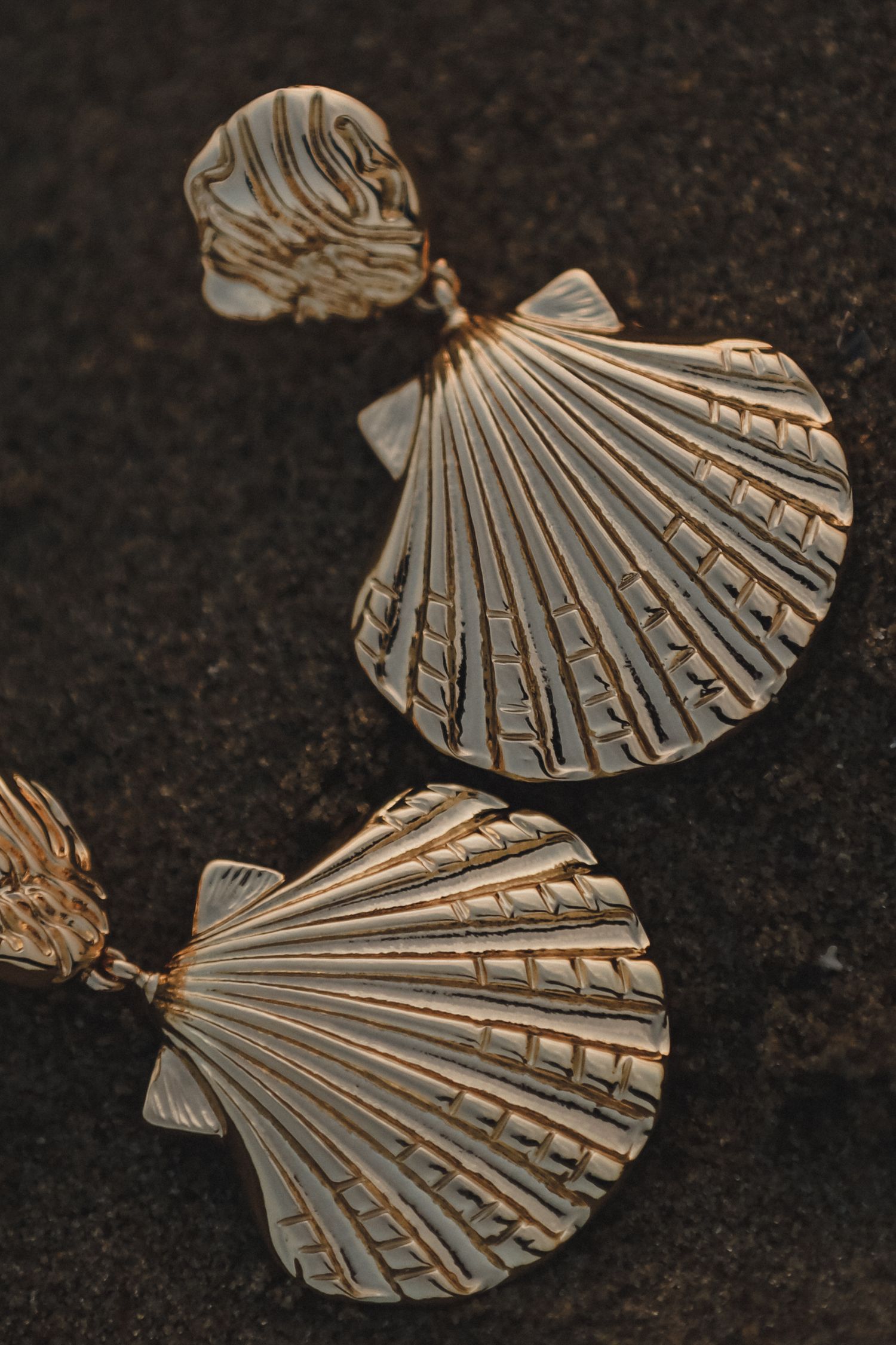 Close-up of gold scallop shell-shaped drop earrings resting on dark textured stone surface.
