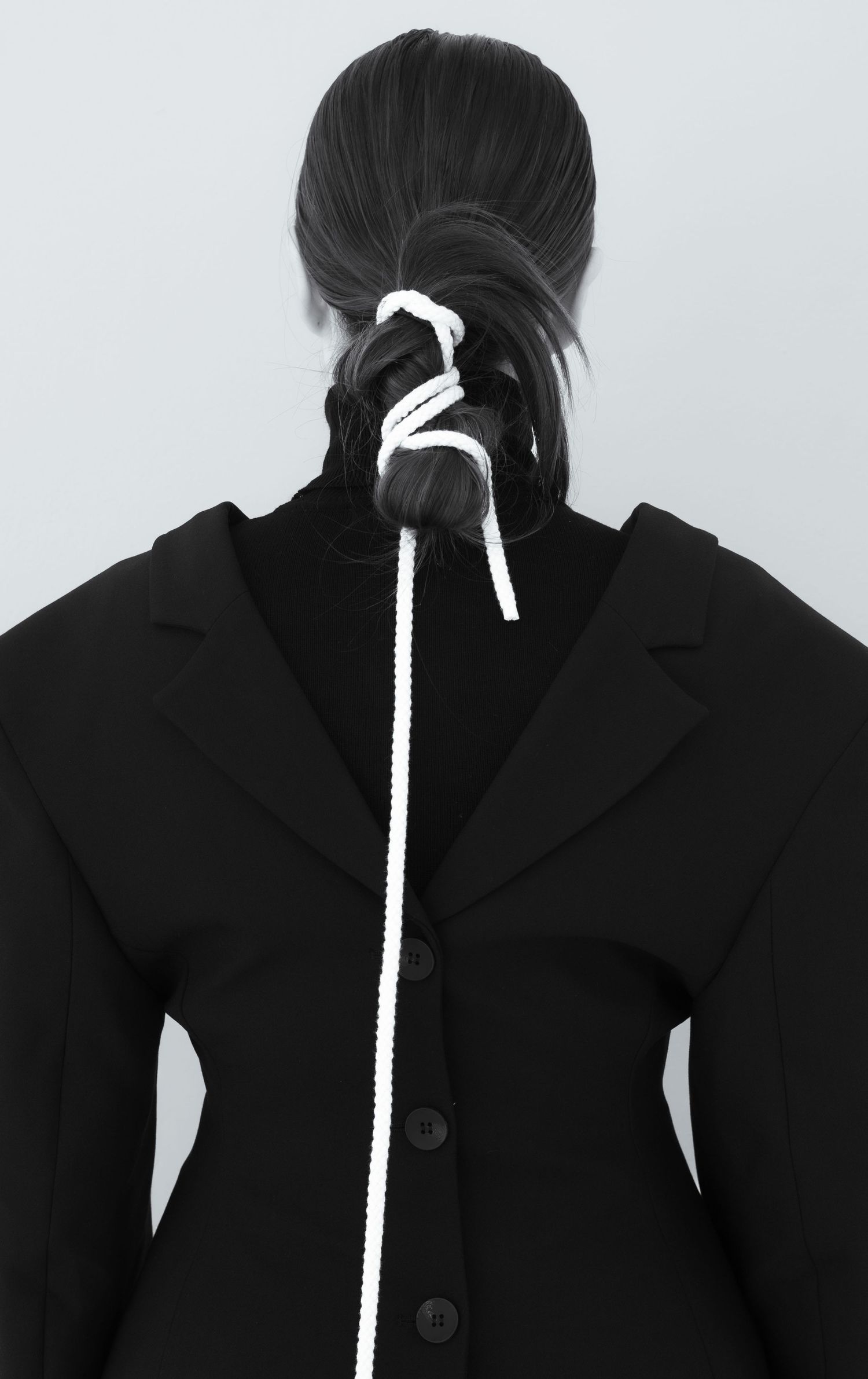 Back view of woman in black blazer with white rope tied through dark hair ponytail.