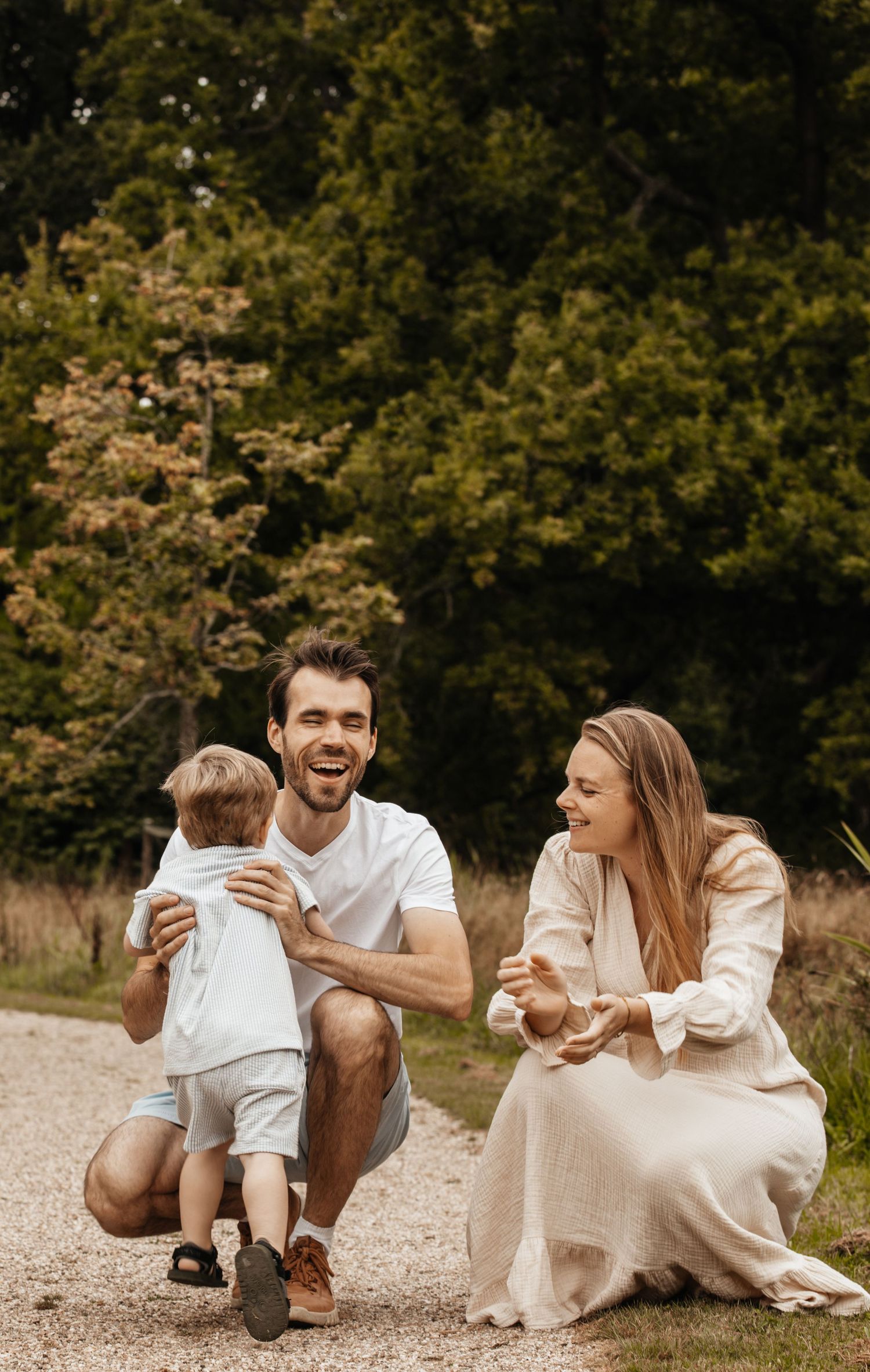 Happy young family with toddler enjoying outdoor walk on gravel path surrounded by lush green trees.