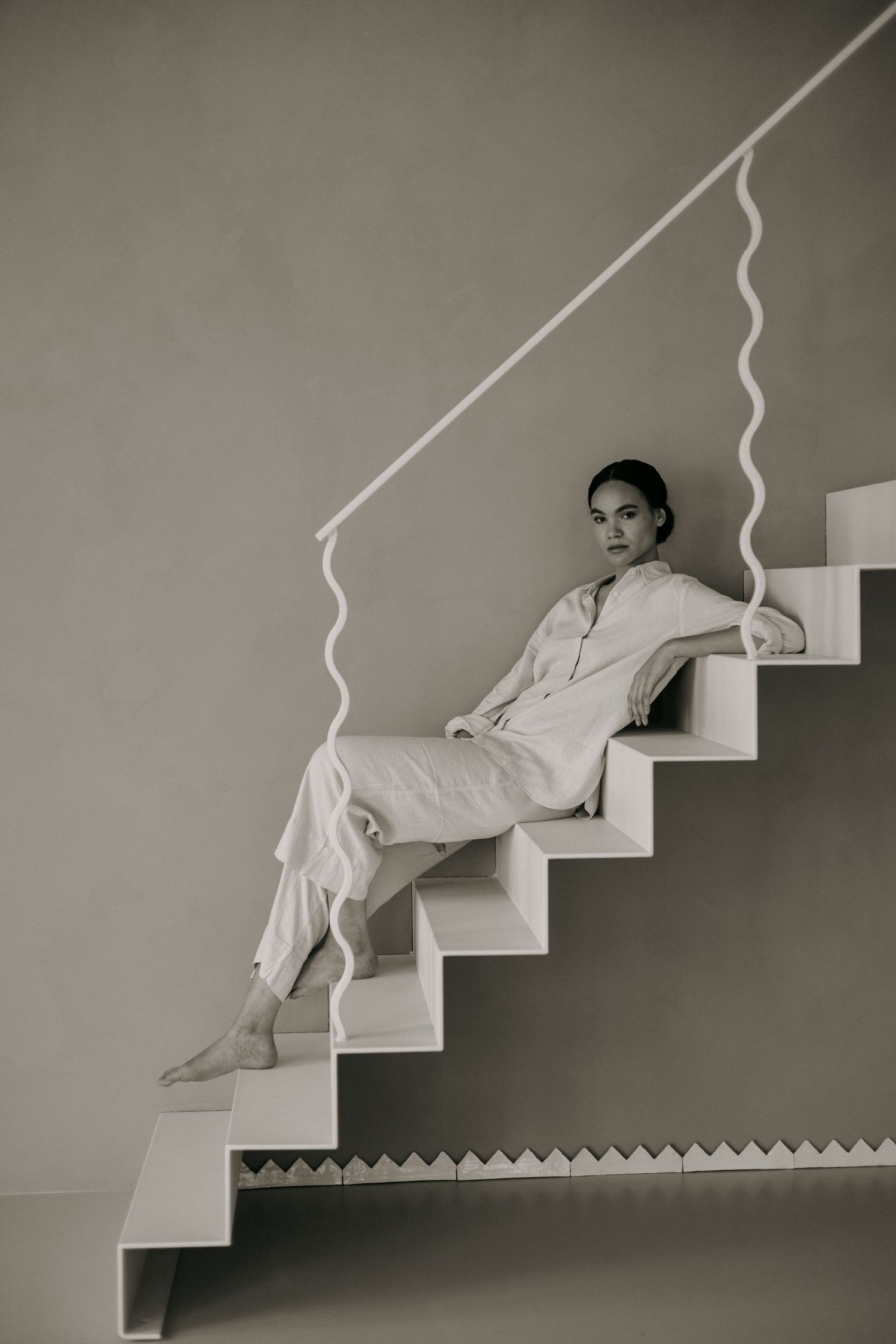 Woman in white linen jumpsuit sitting barefoot on modern white staircase in black and white photo.