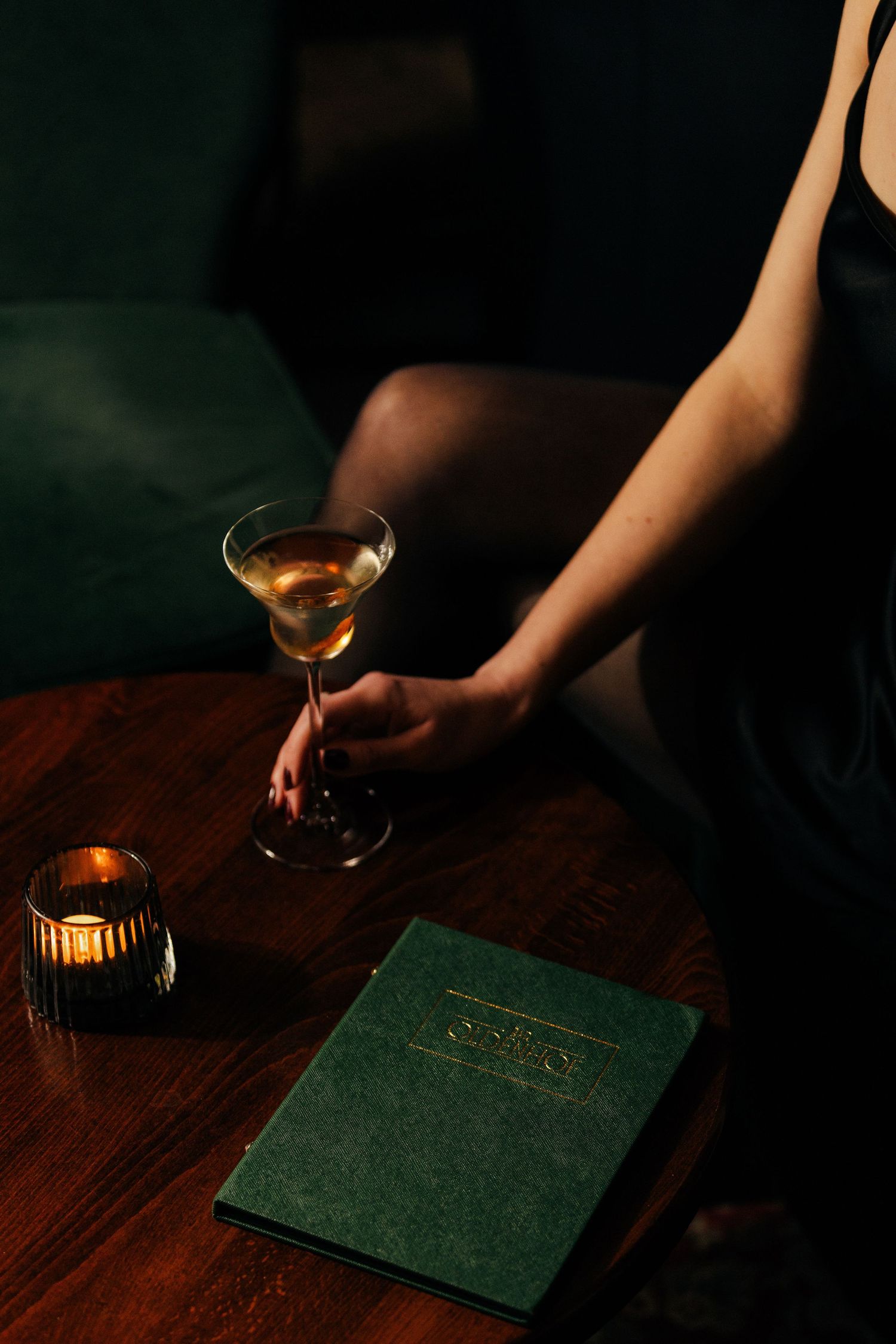 Woman in black dress holding martini glass near candle and green menu book on wooden table.