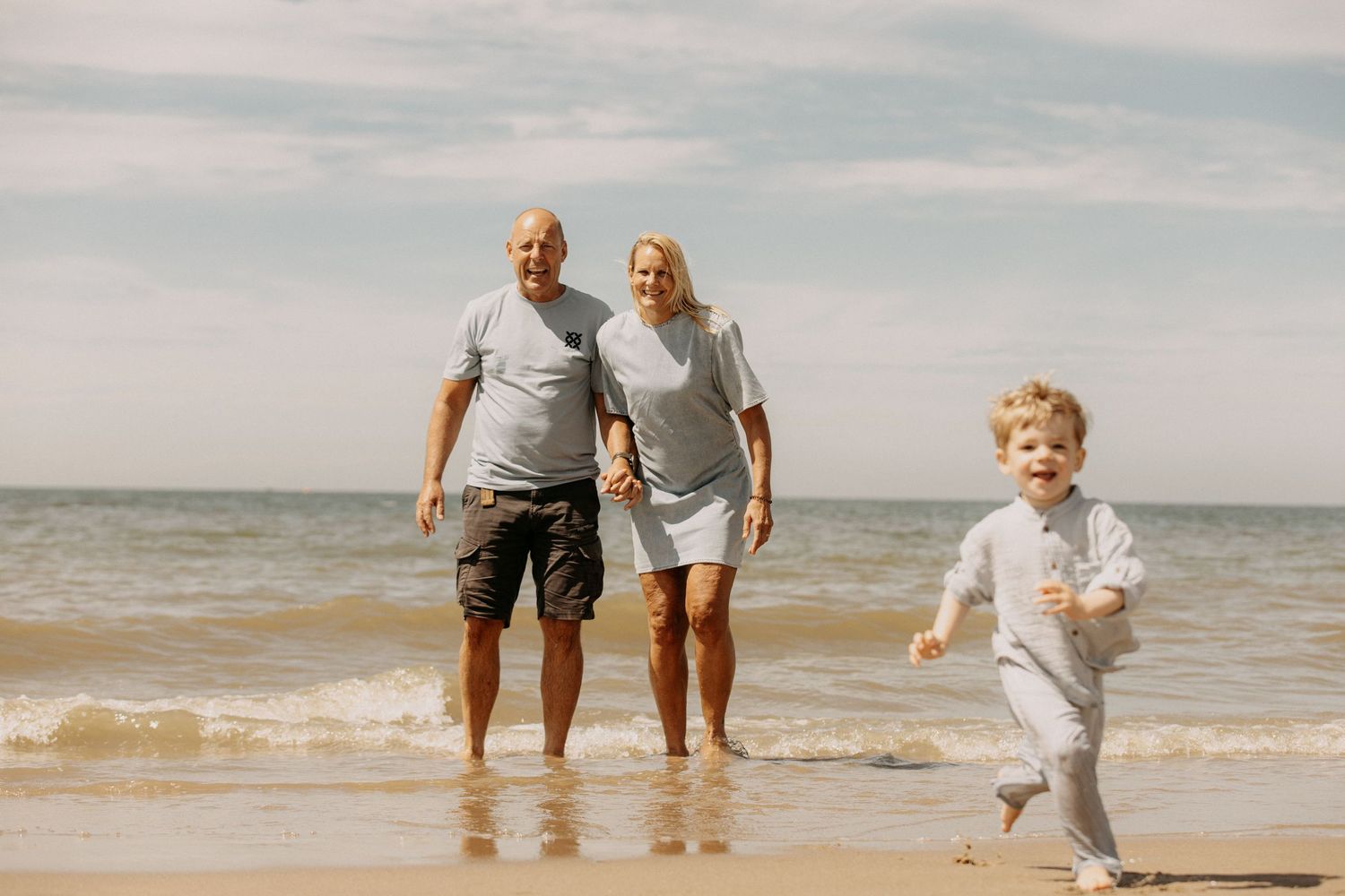Grandparents and toddler enjoying a sunny beach day, with waves and blue sky in the background.