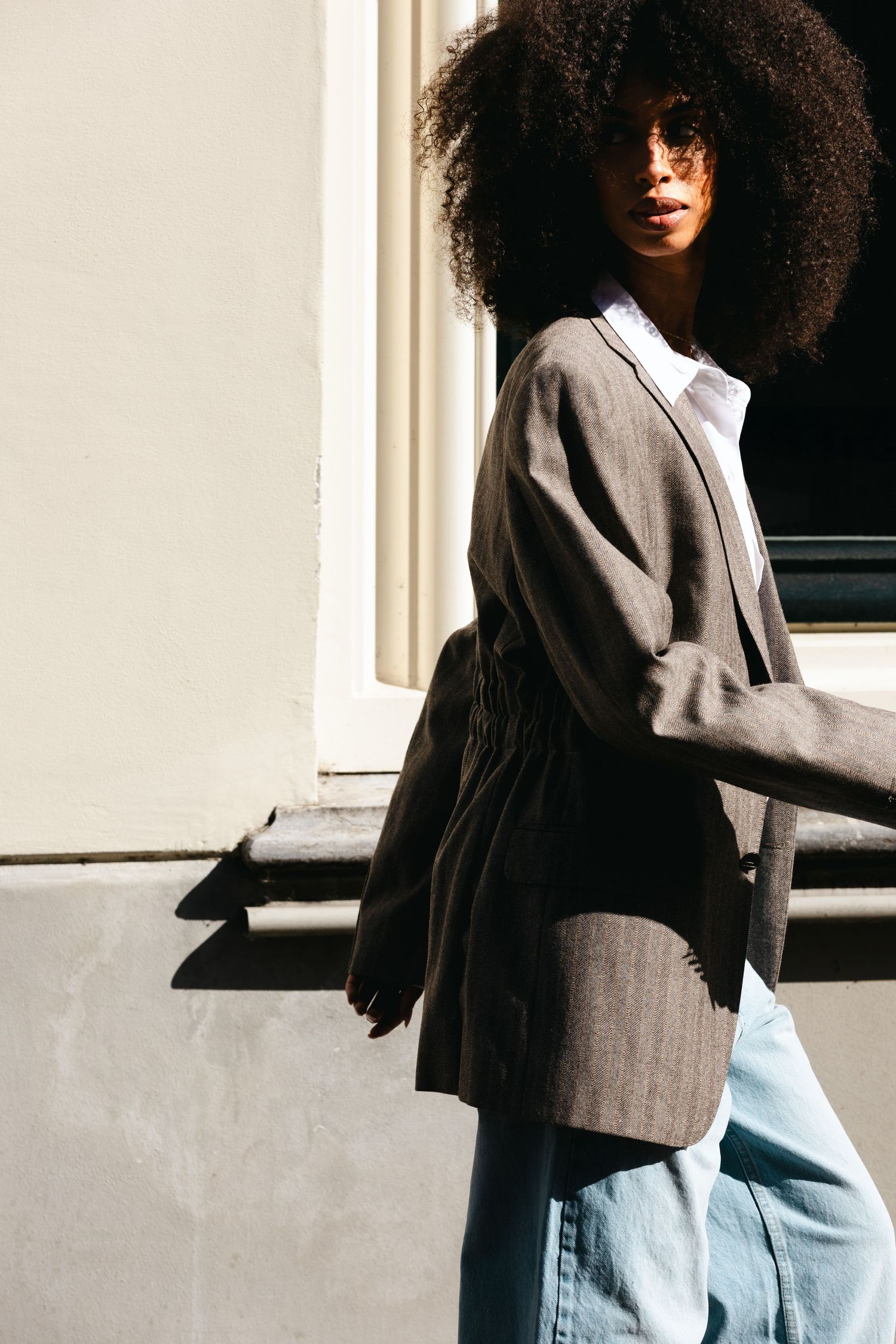 Woman with natural afro hair wearing a gray blazer and white shirt in bright sunlight.