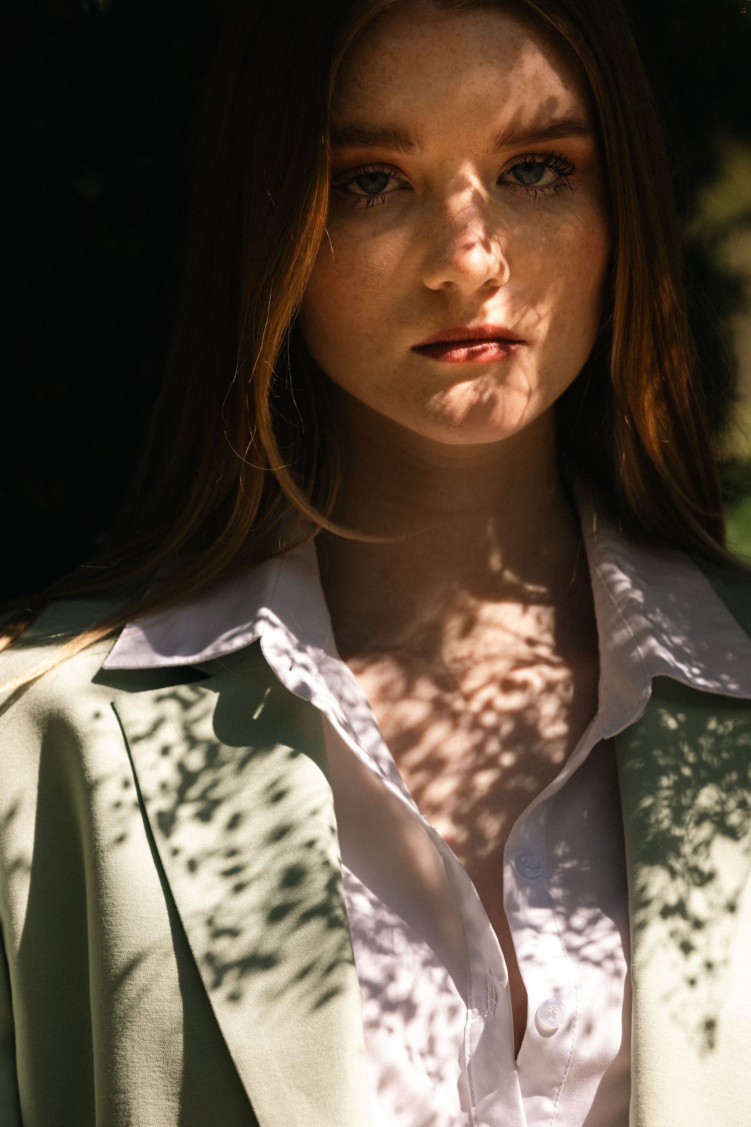 Close-up portrait of freckled blond woman in sage blazer with dappled sunlight.
